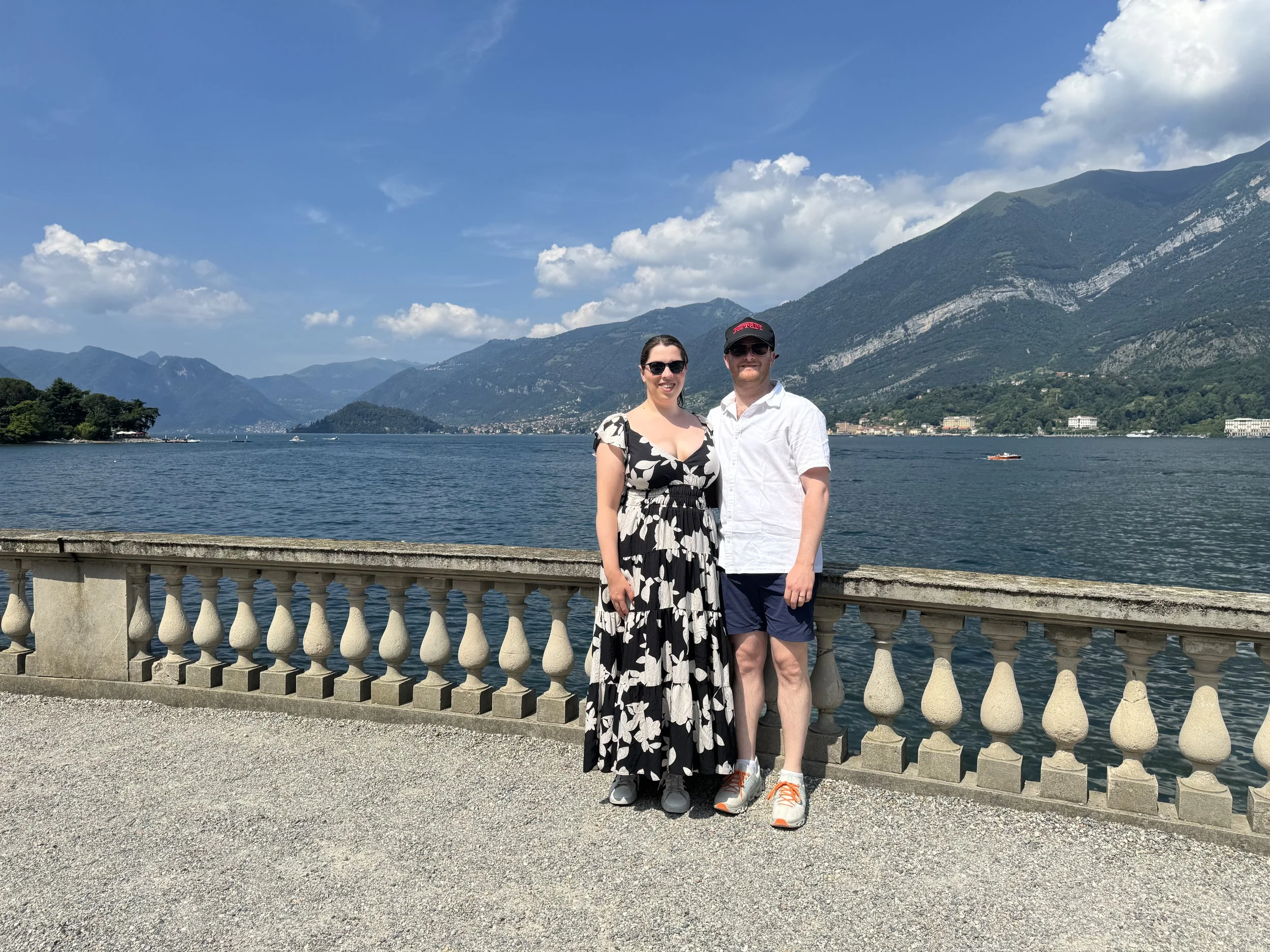 A smiling couple standing side by side by a stone railing overlooking a lake, with mountains and a blue sky with clouds in the background.