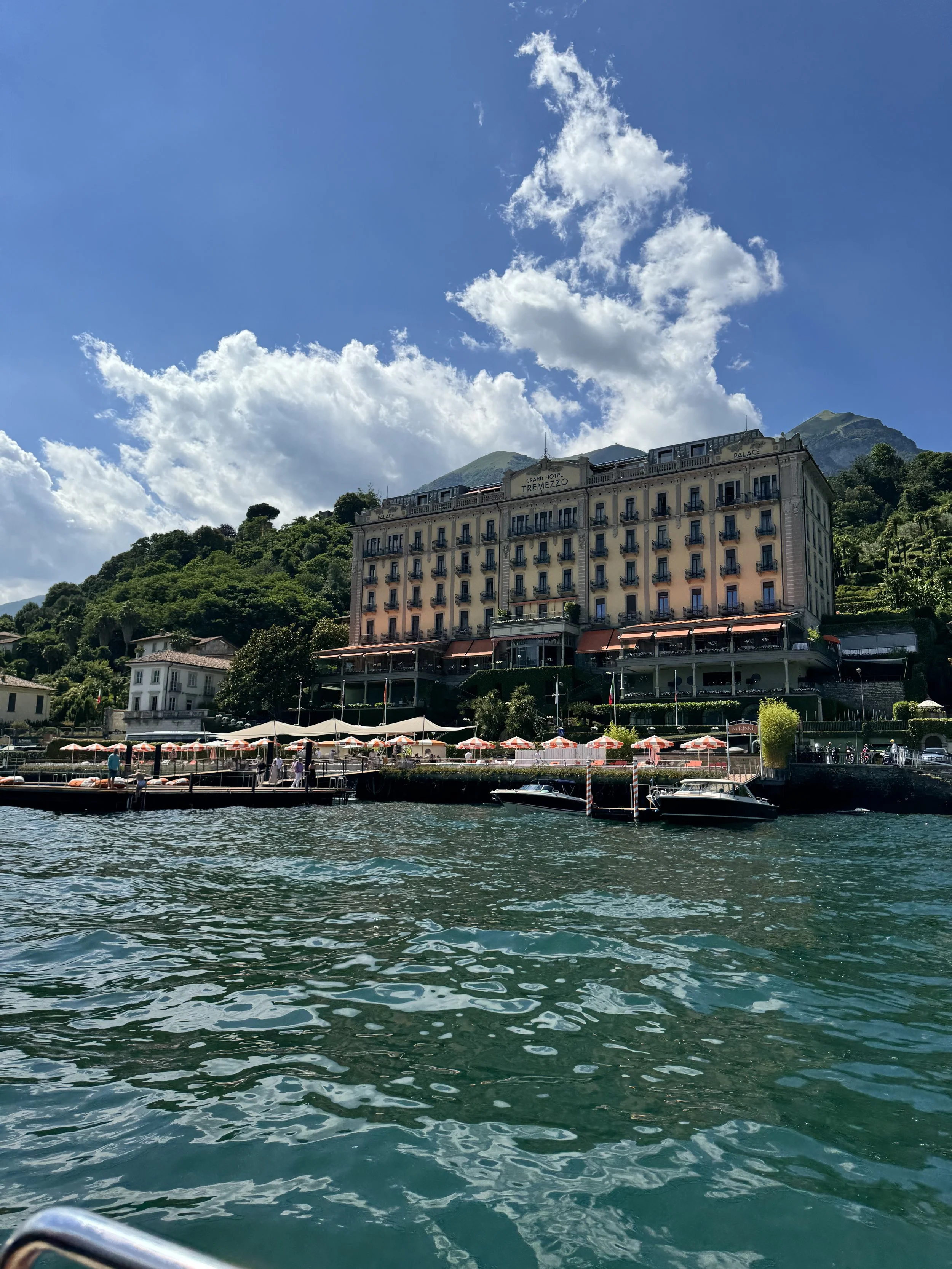 View of a large hotel called Grand Hotel Tremezzo situated along a lake with green hills and mountains in the background. The hotel has multiple balconies and is surrounded by lush greenery. In the foreground, there are deck chairs with umbrellas along the waterfront, and a boat is docked at the pier in the water.