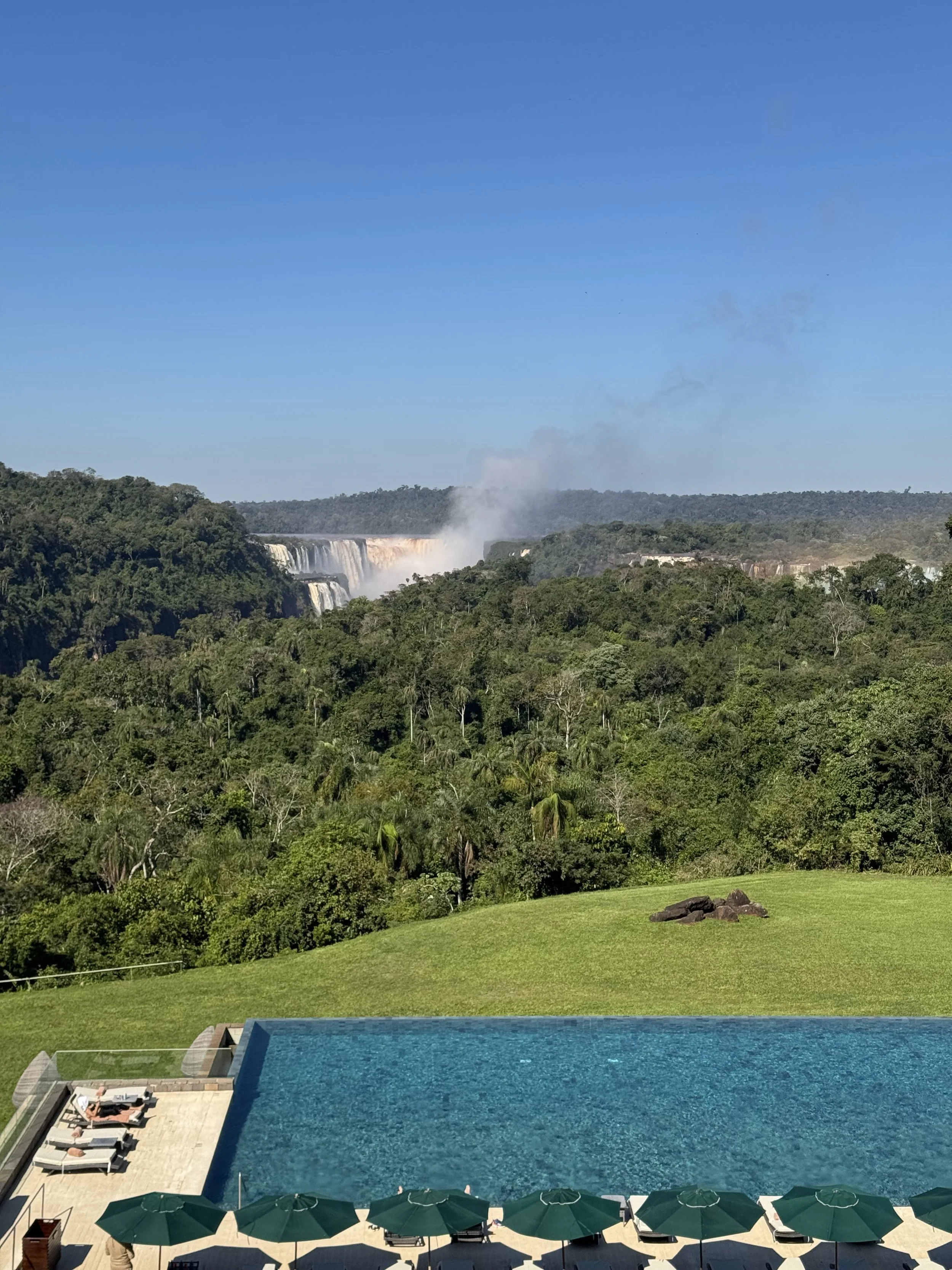A view of a swimming pool with green umbrellas and lounge chairs along its edge, overlooking lush greenery and dense trees. In the background, there are waterfalls and a mist rising from the water, with a bright blue sky overhead.