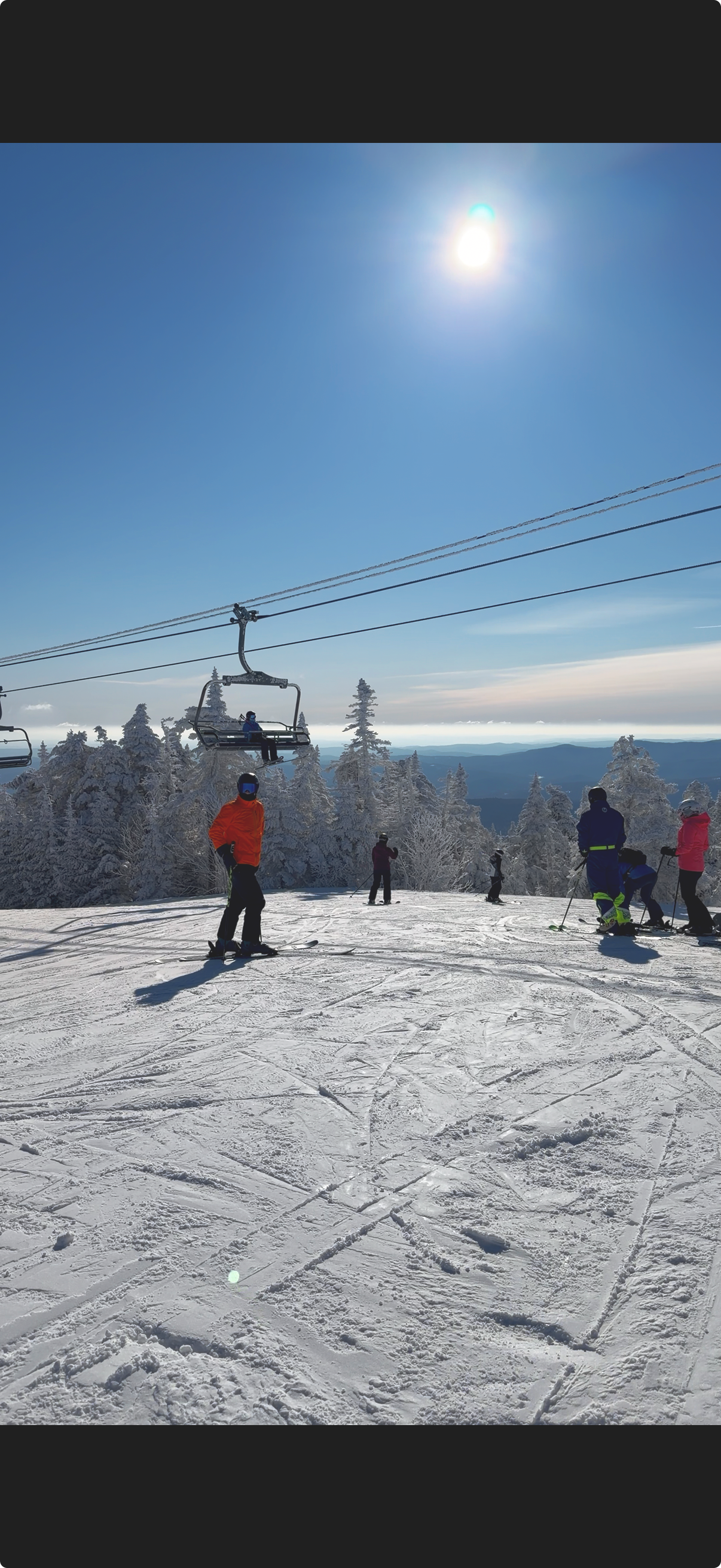 People skiing on a snowy mountain with trees covered in snow, ski lift chairs overhead, bright sun in a clear blue sky.