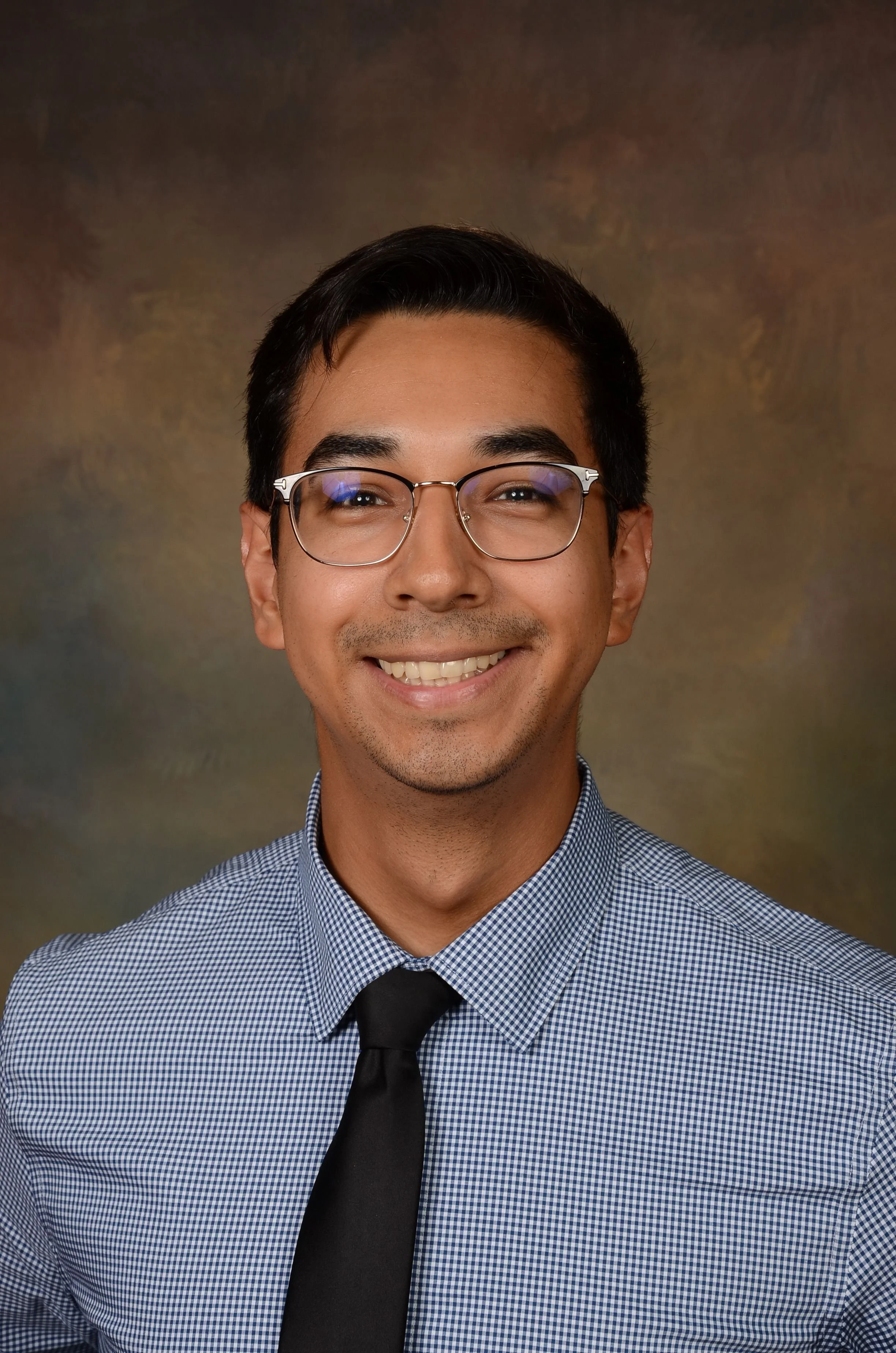 Portrait of a smiling young man with glasses, short dark hair, wearing a blue checkered shirt and black tie, against a brownish studio background.