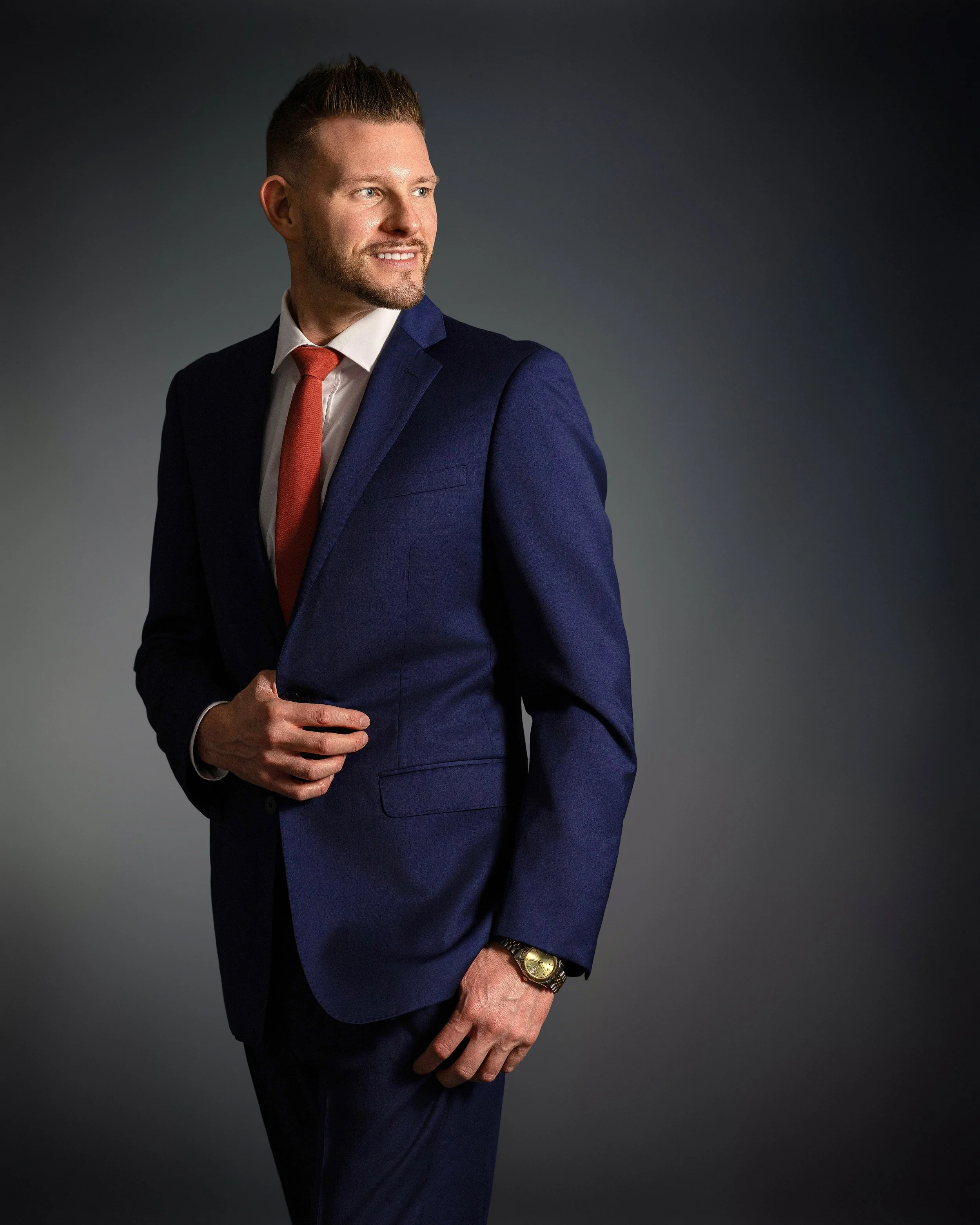 Man in a blue suit with a white shirt and red tie, wearing a gold watch, standing against a dark background.
