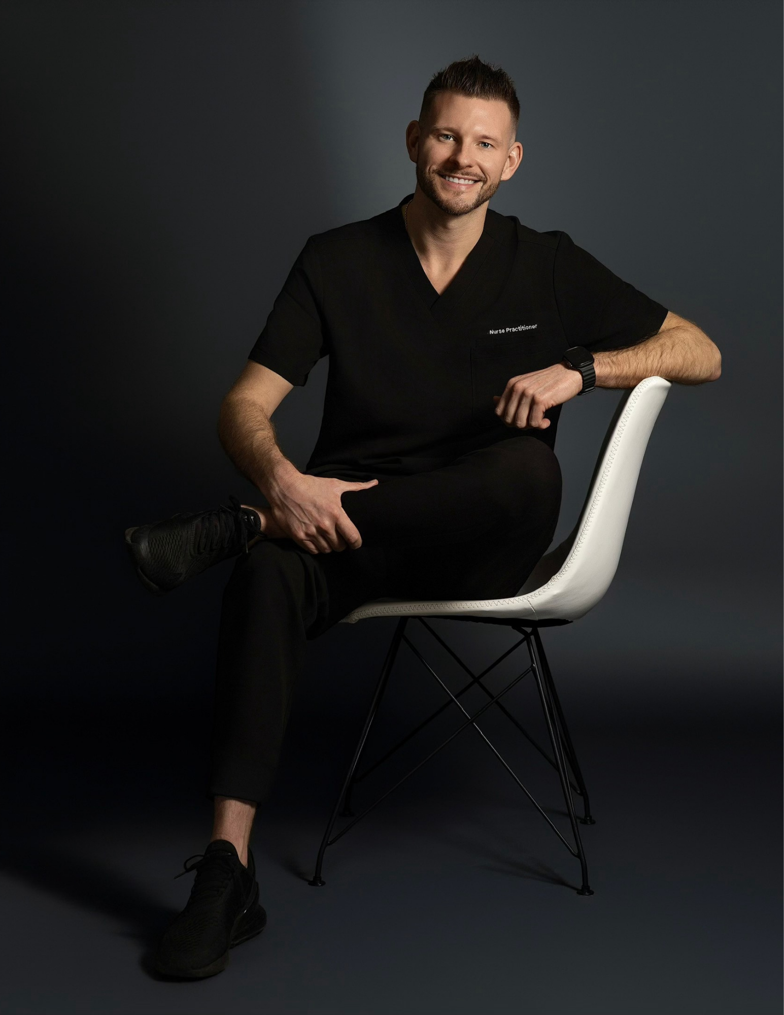 A smiling man in black medical scrubs sits on a white chair against a dark background, with one leg bent and foot resting on the floor, wearing a smartwatch on his left wrist.