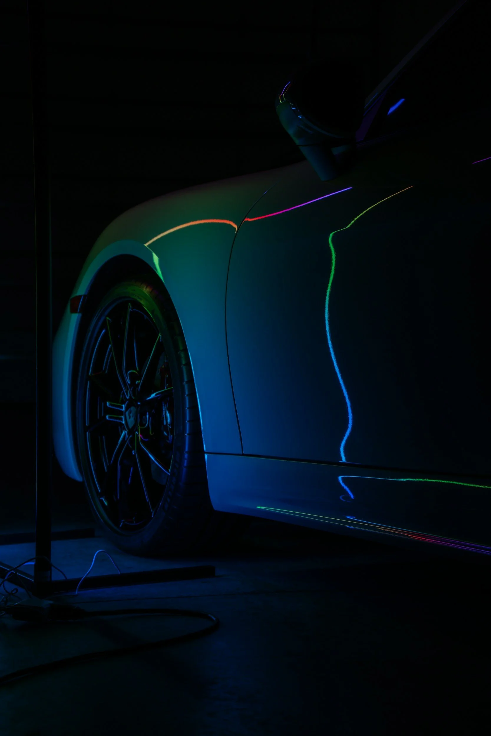 A close-up shot of a modern car with reflections of colorful lights on its surface, set against a dark background.