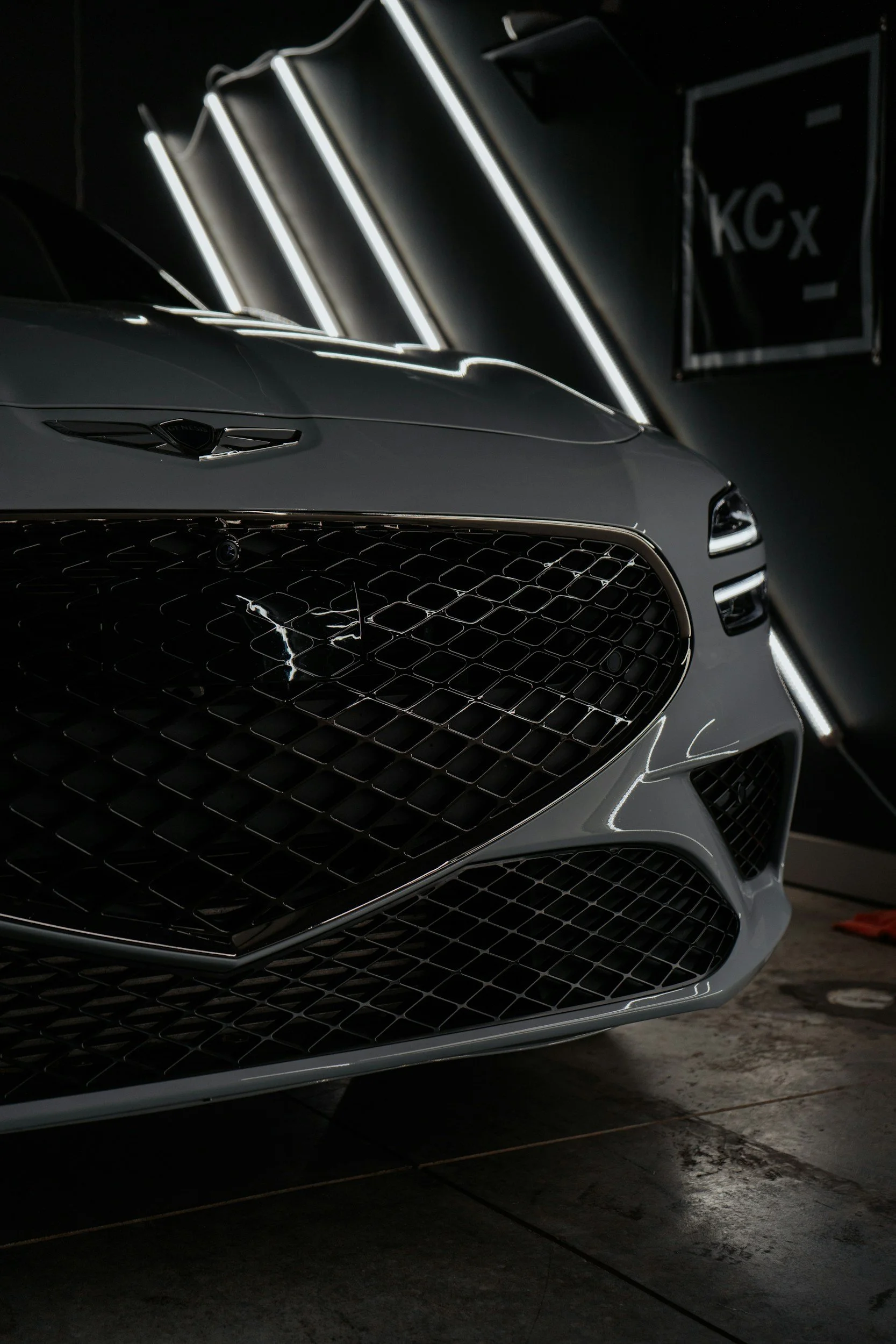 Close-up of the front part of a gray luxury car with a prominent grille, parked indoors with illuminated neon lights in the background.