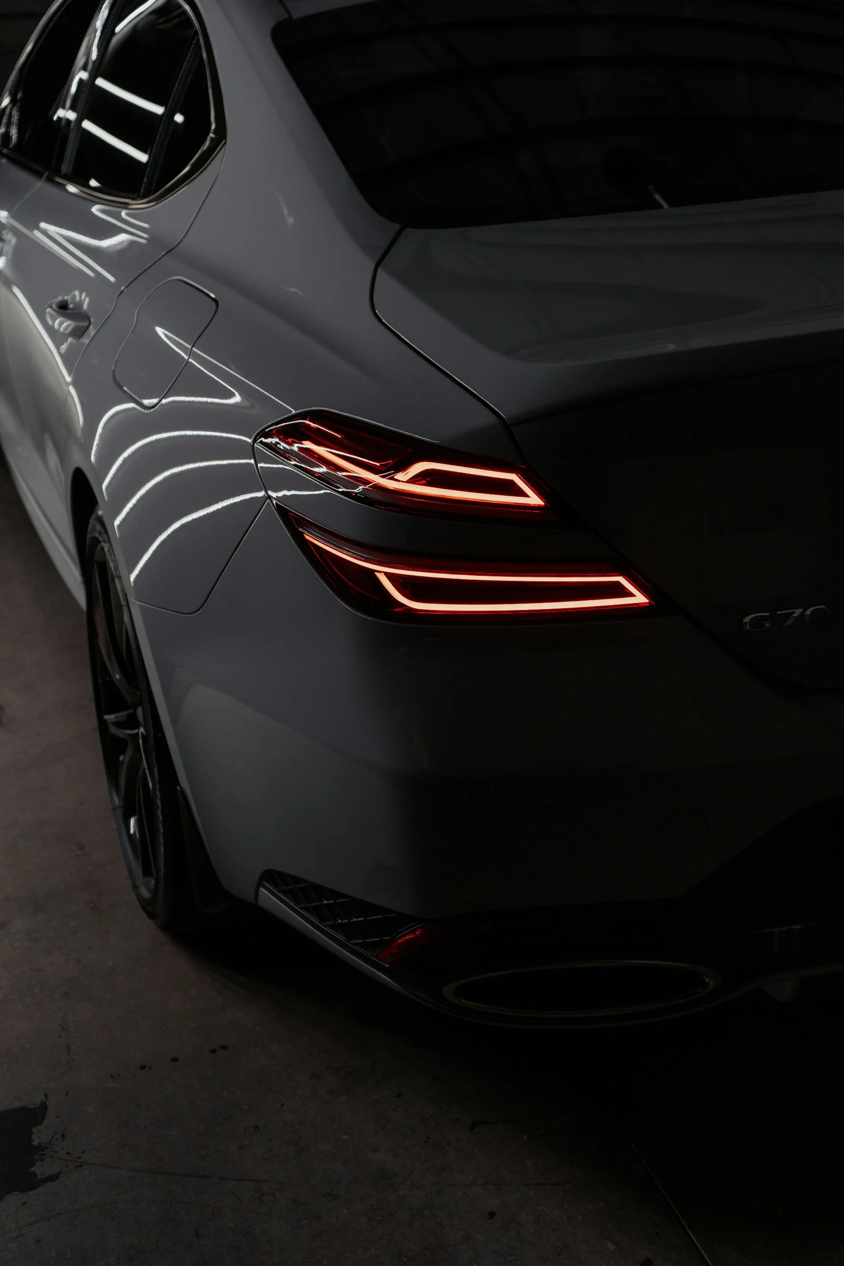 Close-up of the rear and side of a dark gray luxury car in a dimly lit environment. The car's tail lights are illuminated, showing streamlined LED light strips.