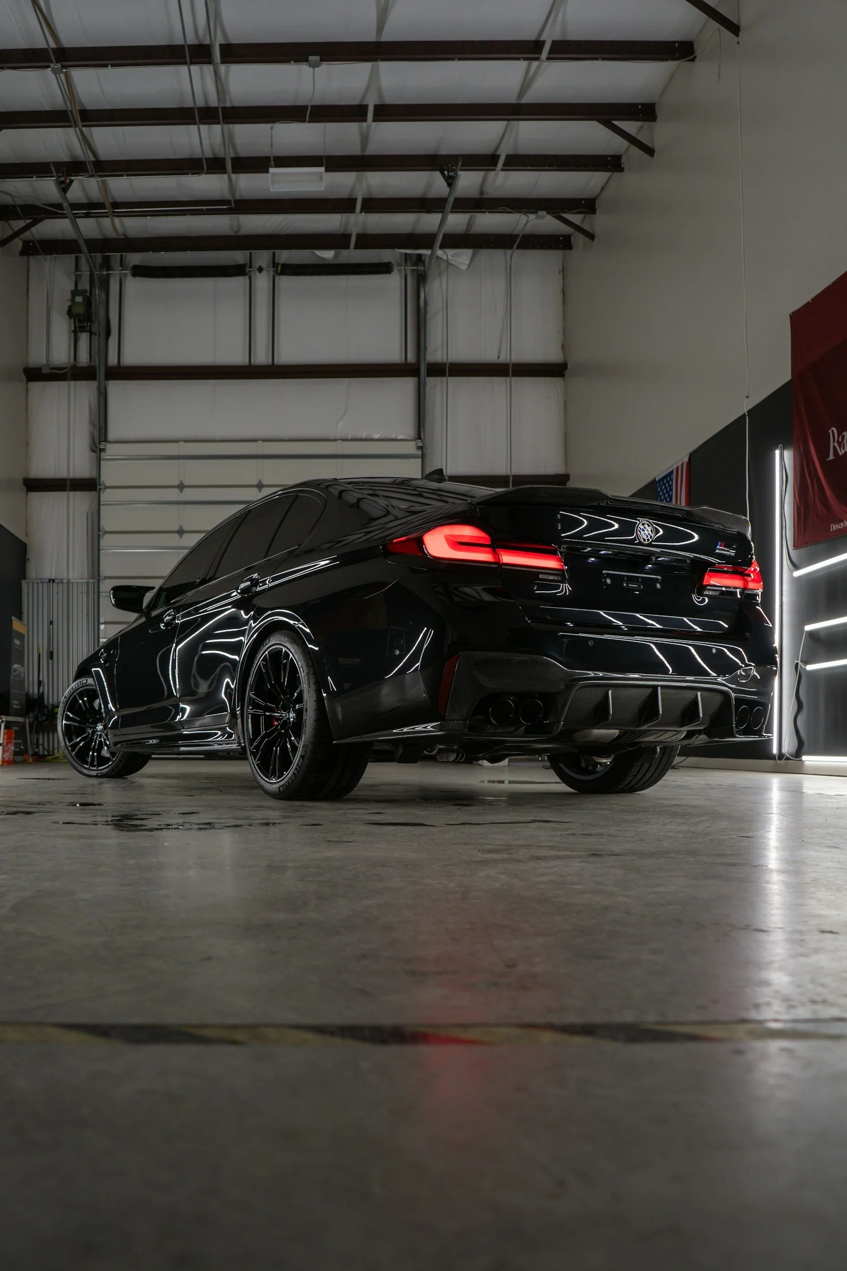 Black sports car parked in a garage with a high ceiling, fluorescent lighting, and plain concrete floor.