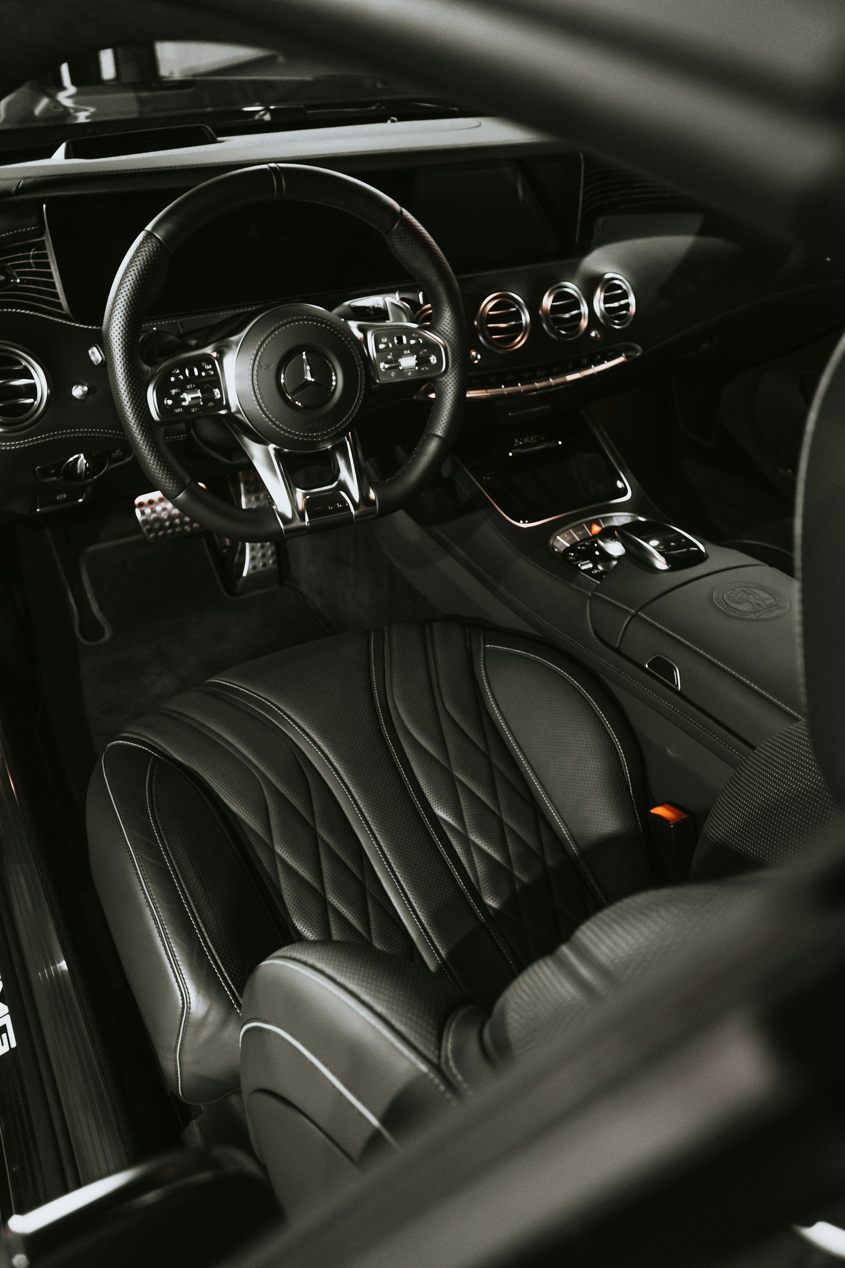 Close-up of the interior of a luxury car, displaying a leather-wrapped steering wheel with a Mercedes-Benz logo, high-tech dashboard, and quilted black leather seats with white stitching.