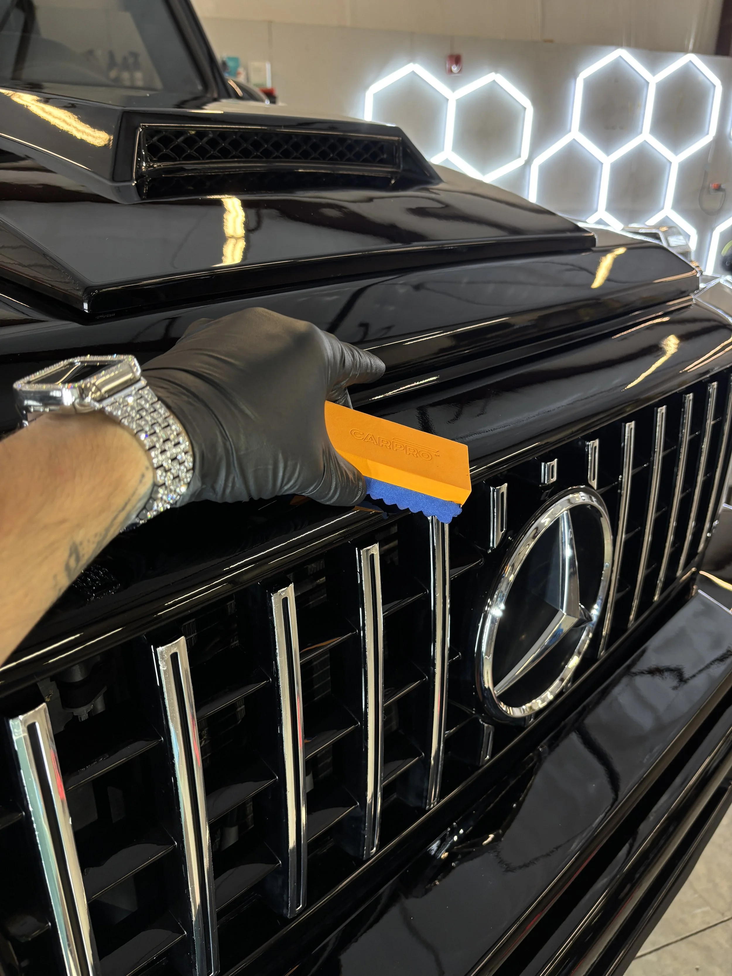 Close-up of a black luxury car with a shiny grille and a hand wearing a black glove holding an orange detailing tool near the front grill. In the background, illuminated hexagonal light fixtures on a wall.