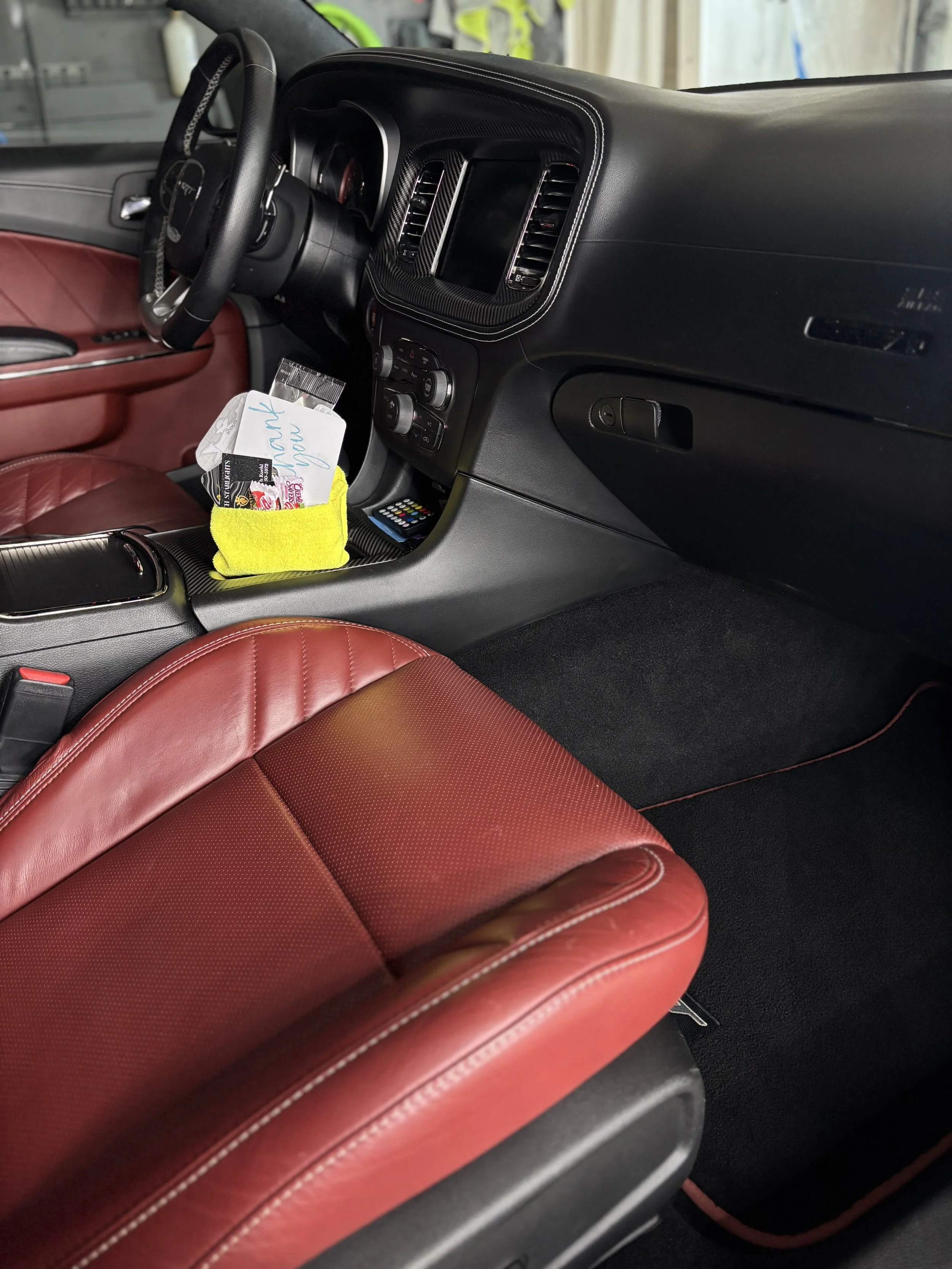 The interior of a car showing red leather seats, a black dashboard with a touchscreen, air vents, and a steering wheel. A yellow cloth with notes and a card are on the center console.