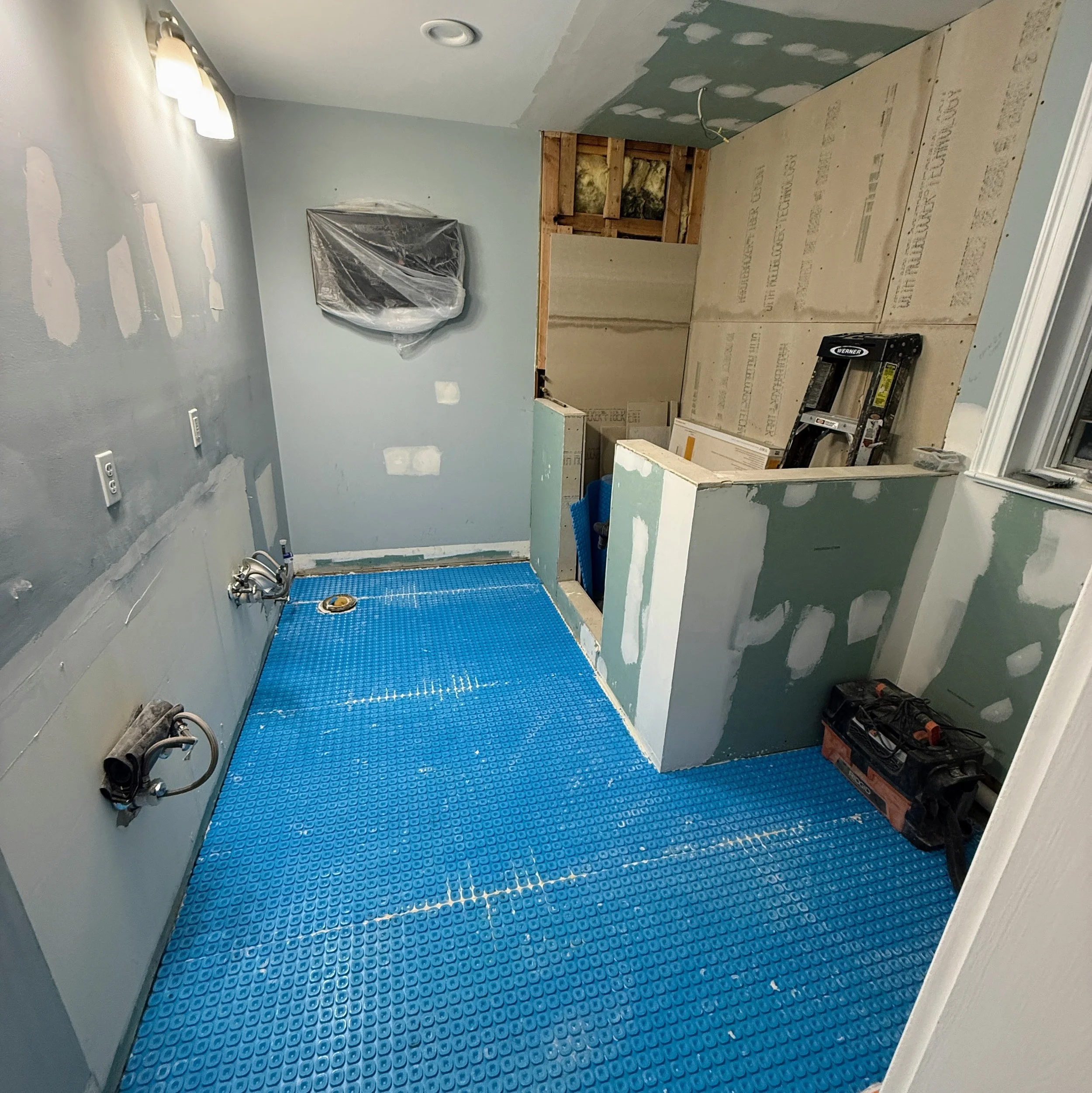 Bathroom under renovation with blue rubber flooring, drywall partially installed, and an unconnected sink with plumbing.