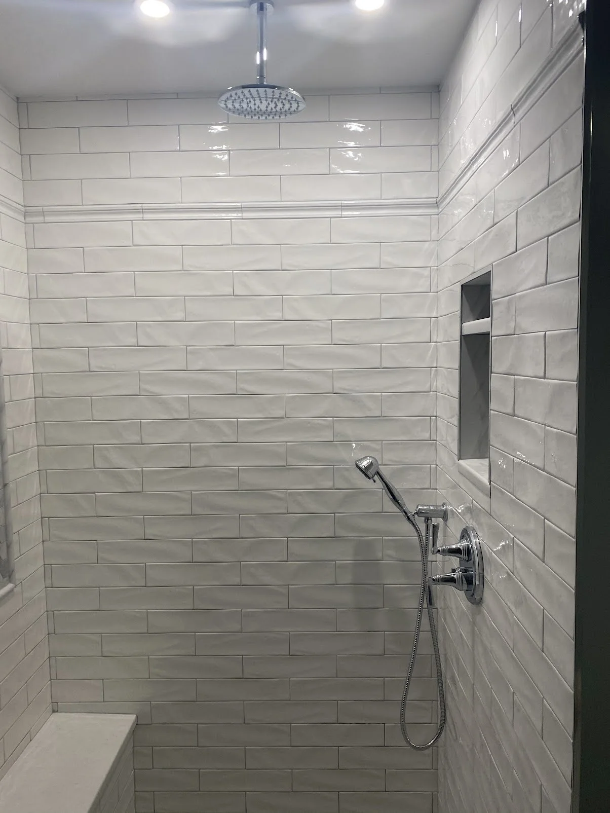 A modern walk-in shower with white subway tile walls, a ceiling-mounted rainfall showerhead, and a handheld showerhead with controls. There's a built-in niche for toiletries.