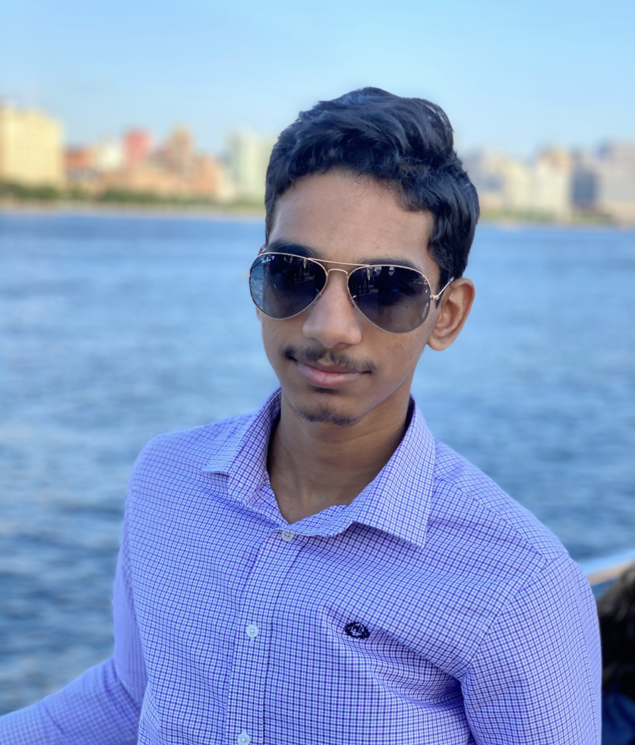 Young man with short dark hair wearing aviator sunglasses and a purple checkered shirt, standing near water with a city skyline in the background.