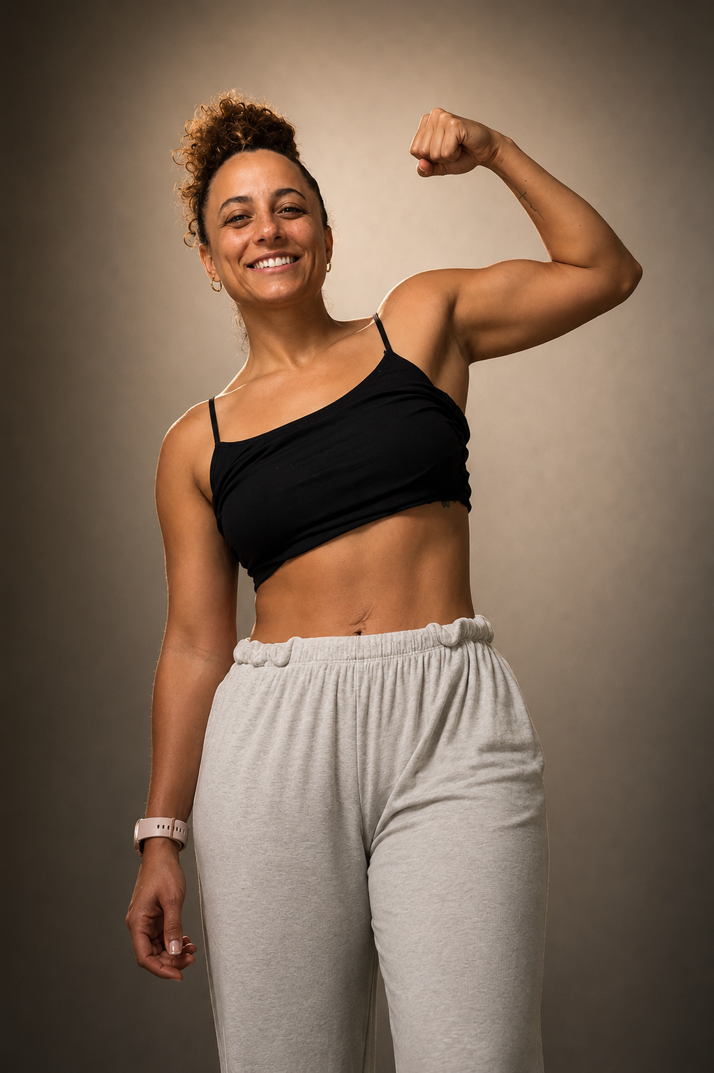 A woman flexing her bicep and smiling, wearing a black crop top, beige sweatpants, and a watch, against a plain background.