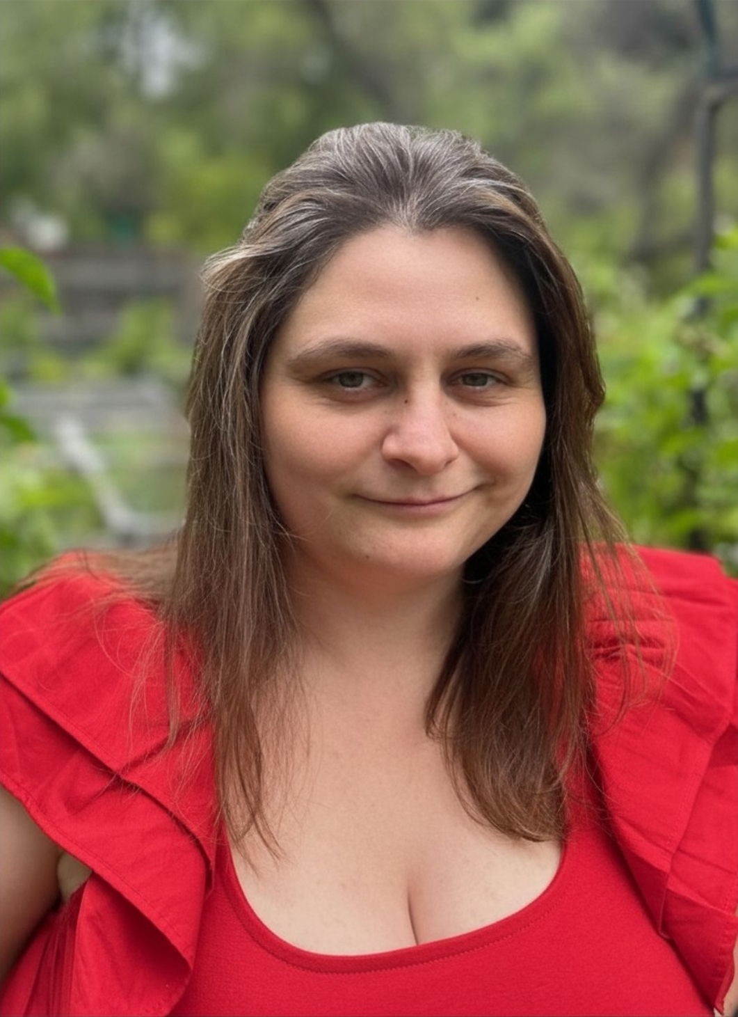 A woman with brown hair and fair skin smiling outdoors, wearing a red top, with greenery in the background.