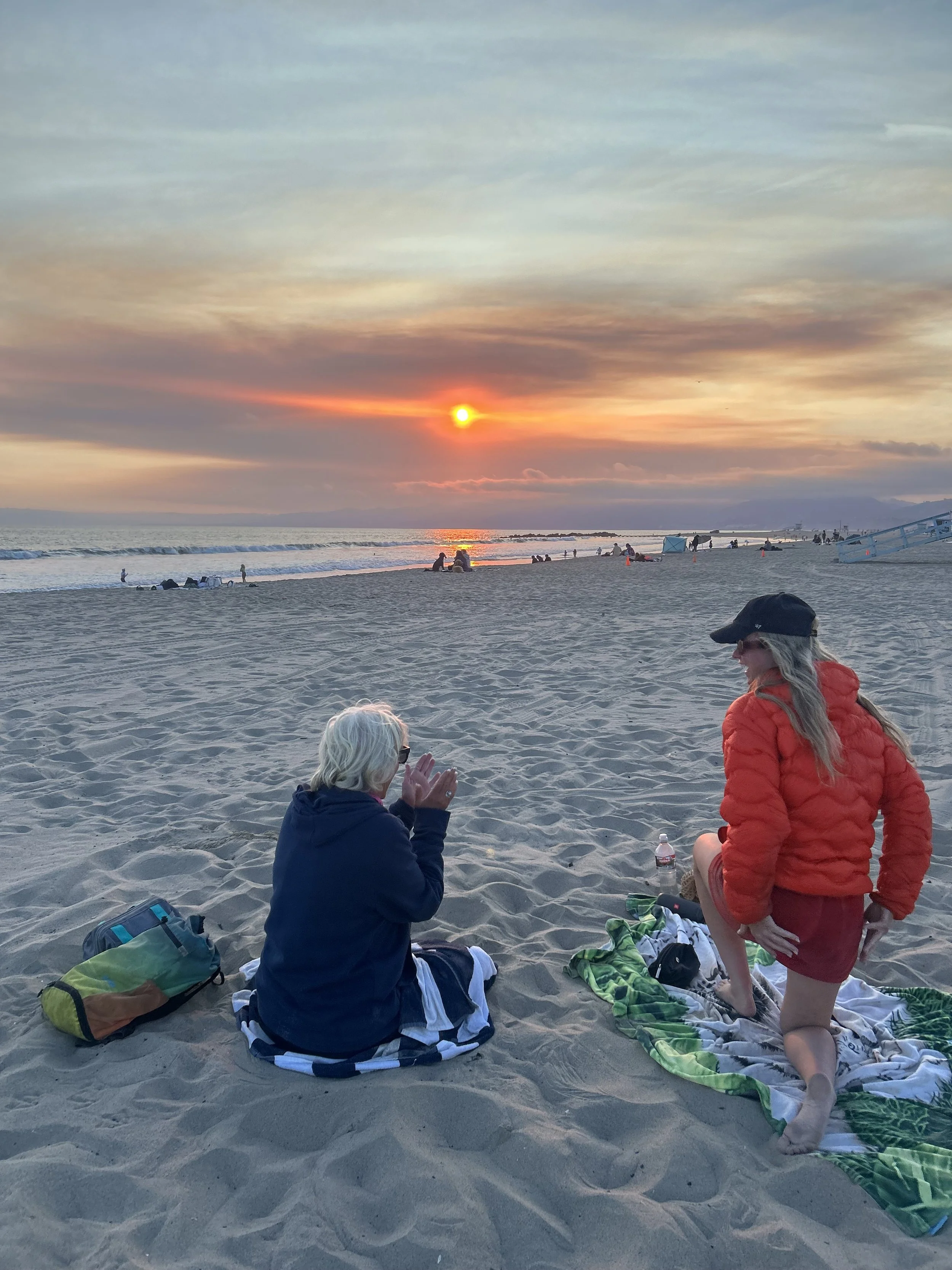 A sunset at the beach with people sitting and lying on the sand, some chatting, some watching the sunset, and others walking near the water. The image shows a serene beach scene with scattered beach items and a colorful sky.