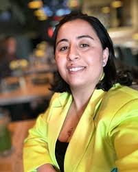 A woman with shoulder-length black hair, wearing a bright yellow blazer, smiling at the camera in an indoor setting.