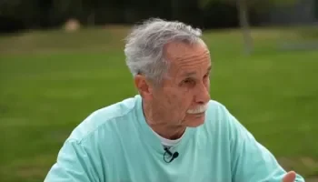 Older man with gray hair and mustache wearing a light blue shirt sitting outdoors on a grassy area.