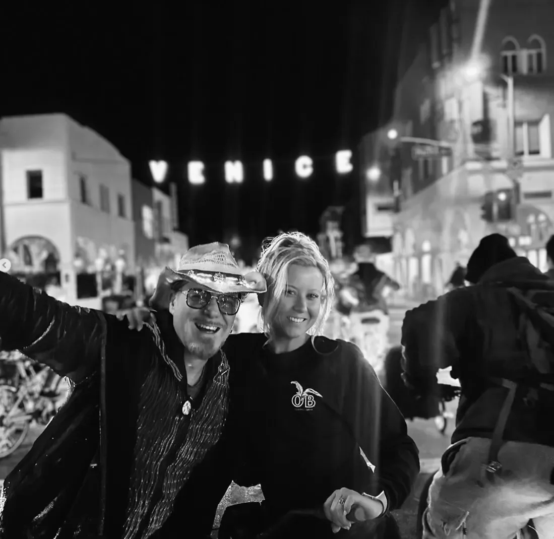 Two people smiling at night in an outdoor area with buildings and a 'Venice' sign in the background.