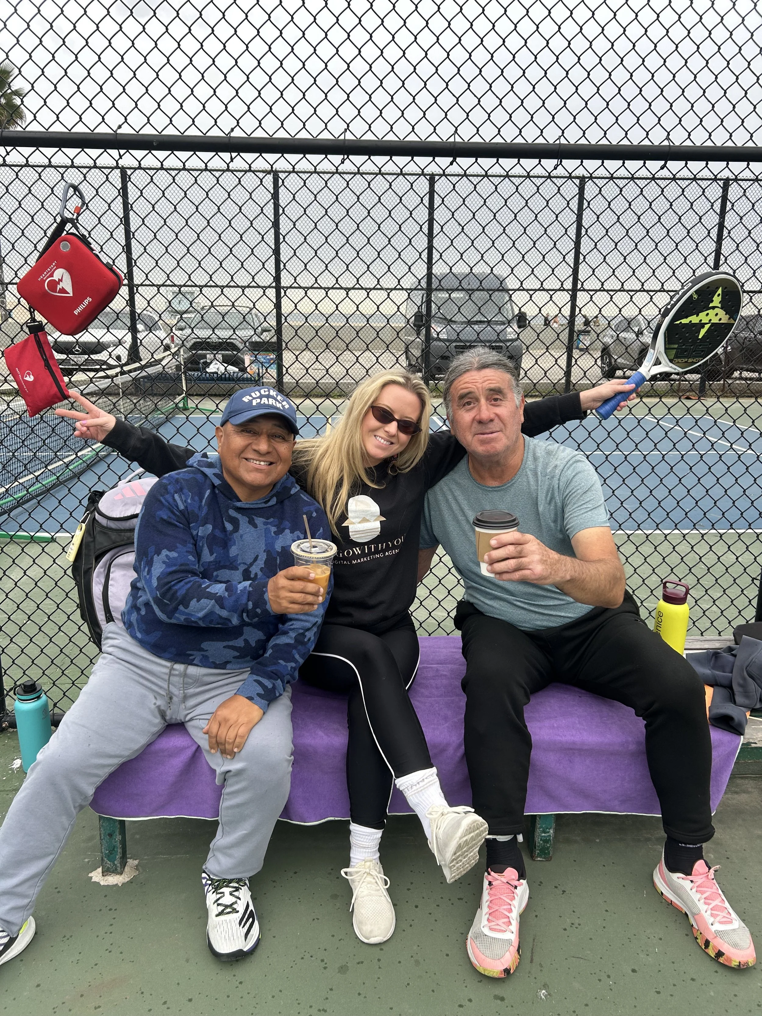 Three people sitting on a tennis court bench behind a chain-link fence, holding drinks, with tennis rackets and bags nearby, in an outdoor setting.