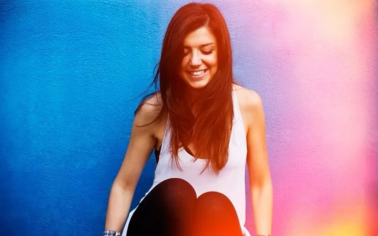 A young woman with long red hair smiling, sitting against a bright blue wall, wearing a white tank top.