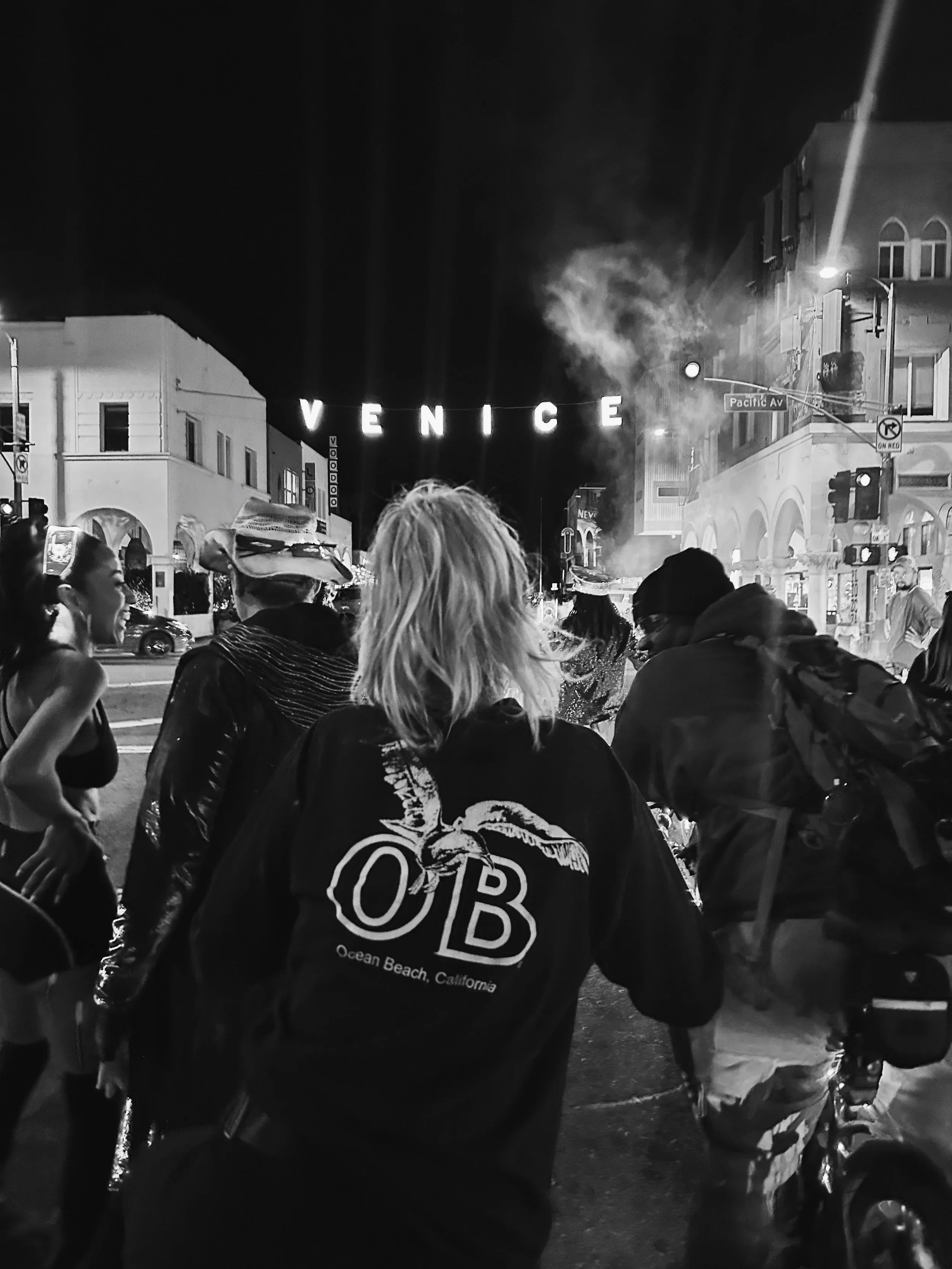 Nighttime street scene in Venice, California, with a crowd of people and a large sign that reads 'VENICE'.