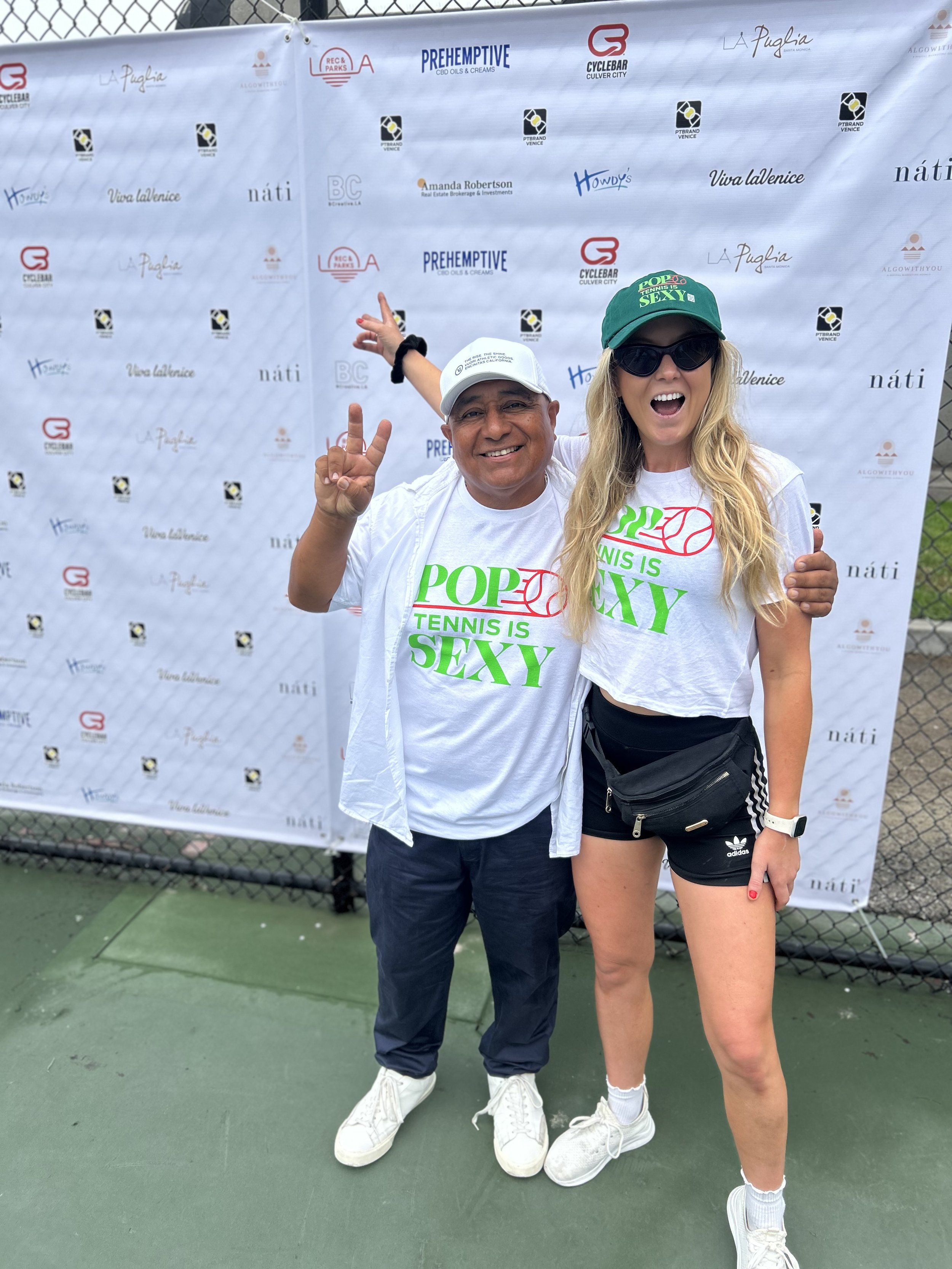 Two people, a man and a woman, standing on a tennis court in front of a step and repeat banner with various sponsor logos. The man is smiling and making a peace sign with his fingers. The woman is wearing sunglasses, a green cap that says 'POPS TENNIS IS SEXY', and is laughing with her mouth open. The man is wearing a dark jacket, white shirt, white shoes, and a white cap. The woman is wearing a white t-shirt with green and red text, black shorts with white stripes, and a white watch.