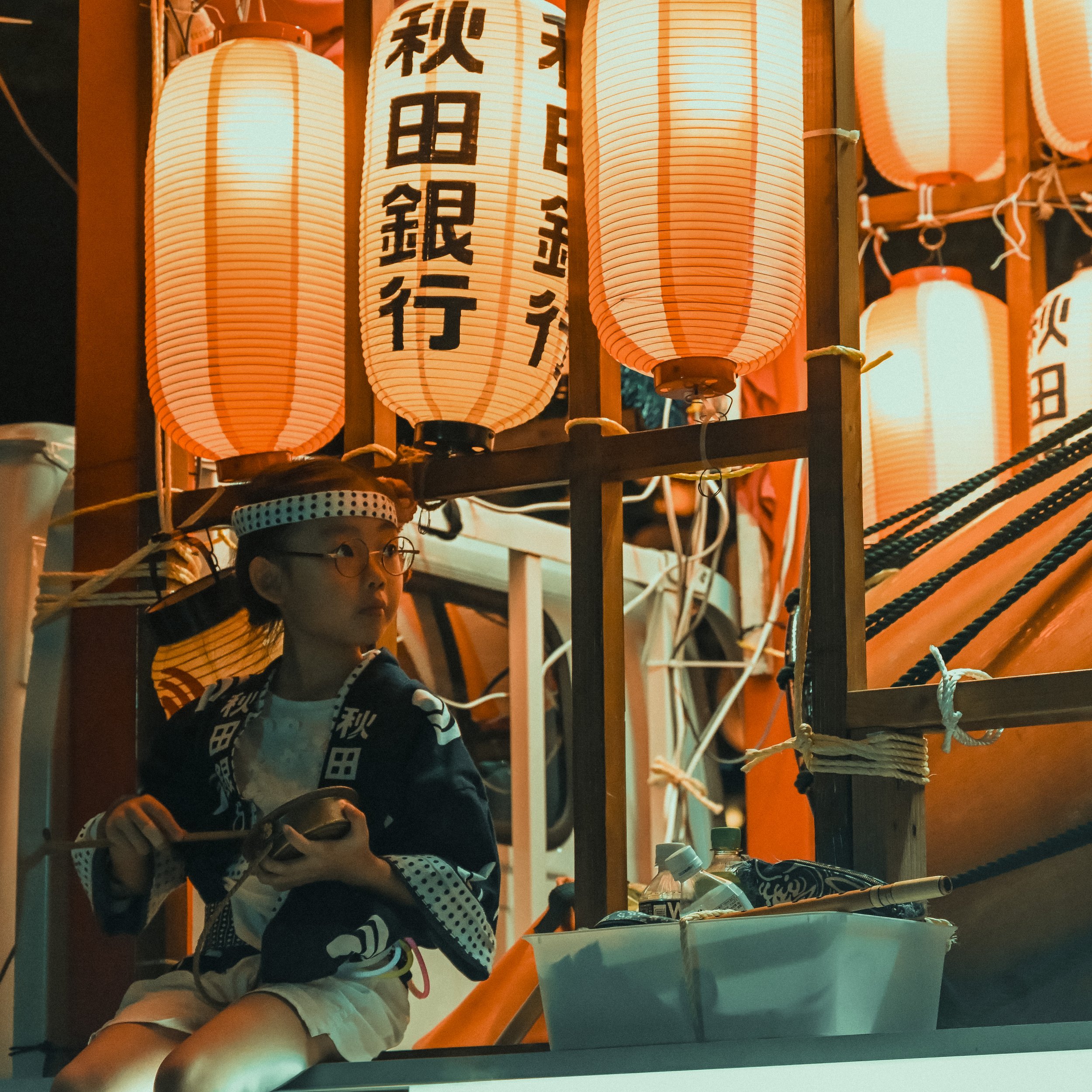 A young person wearing glasses and a patterned hoodie, sitting at a table with various objects including a water bottle, with a display of hanging paper lanterns with Japanese characters illuminated behind them.