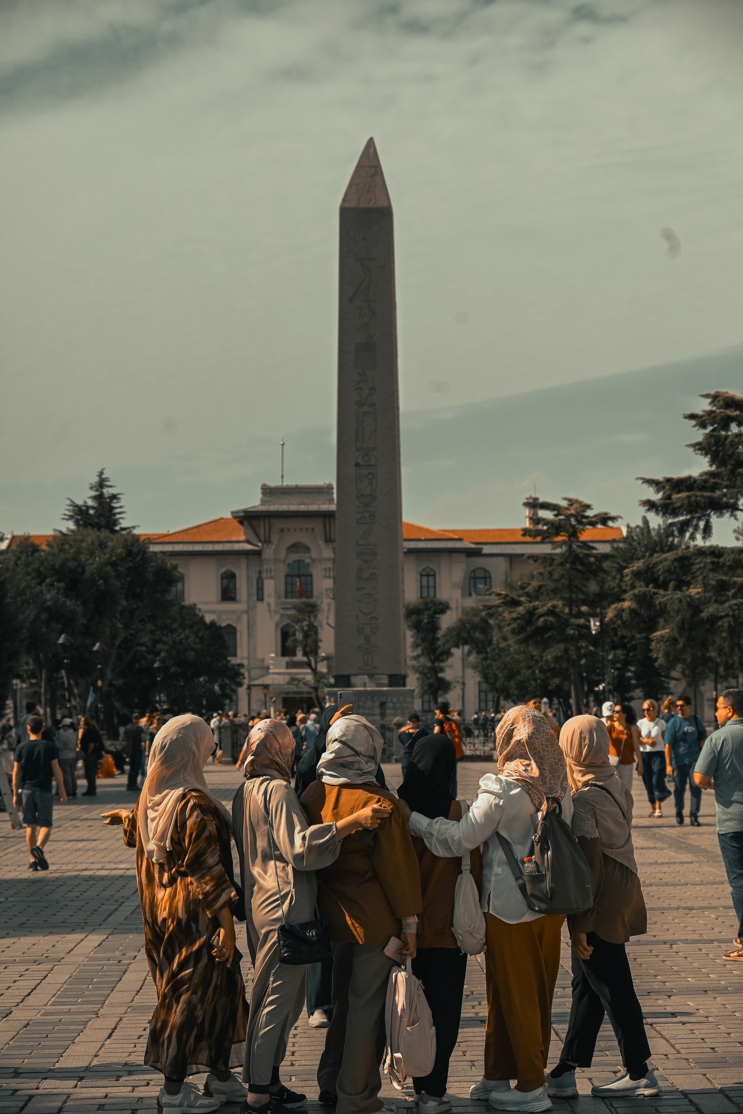 Group of women in hijabs and casual clothing holding hands and standing together in a square surrounded by trees, with a large monument and a historic building in the background and a cloudy sky above.