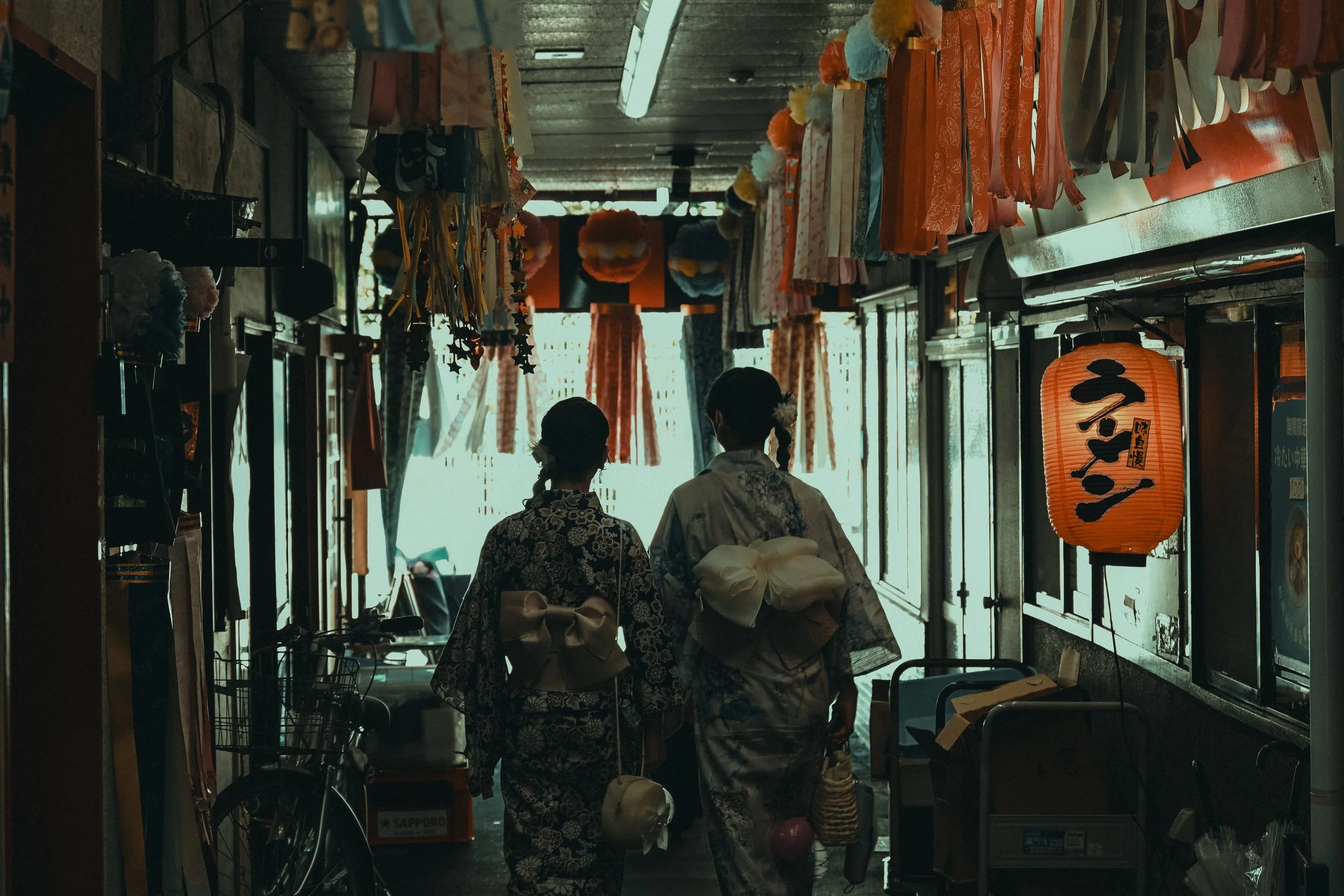Two women in traditional Japanese kimonos walking inside a shop with various colorful items hanging from the ceiling, illuminated by natural light coming from the entrance.