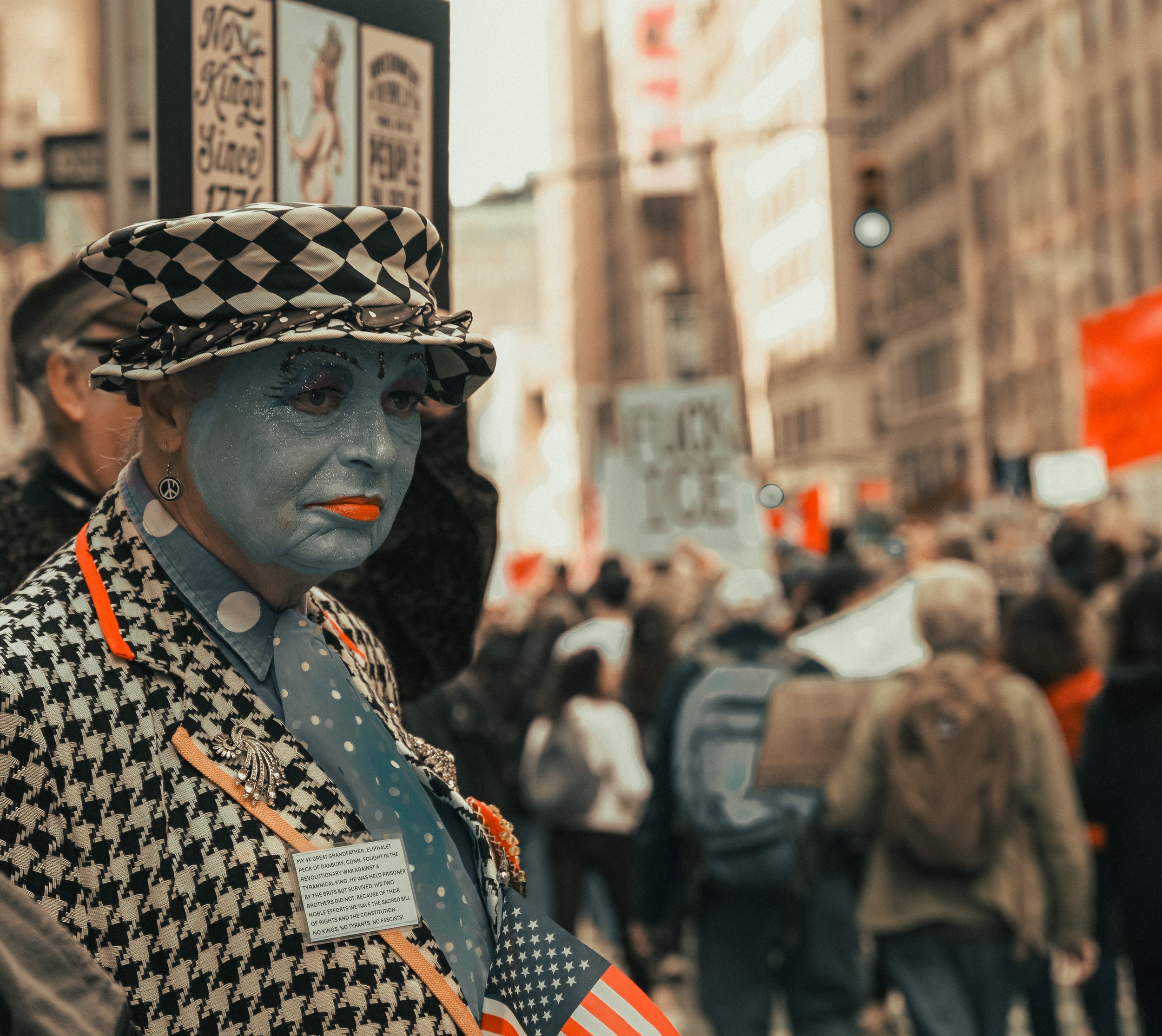 Close-up of an elderly woman with painted blue face, wearing a black and white checkered hat, earrings, and a patterned jacket, standing in a store with shelves of products in the background.