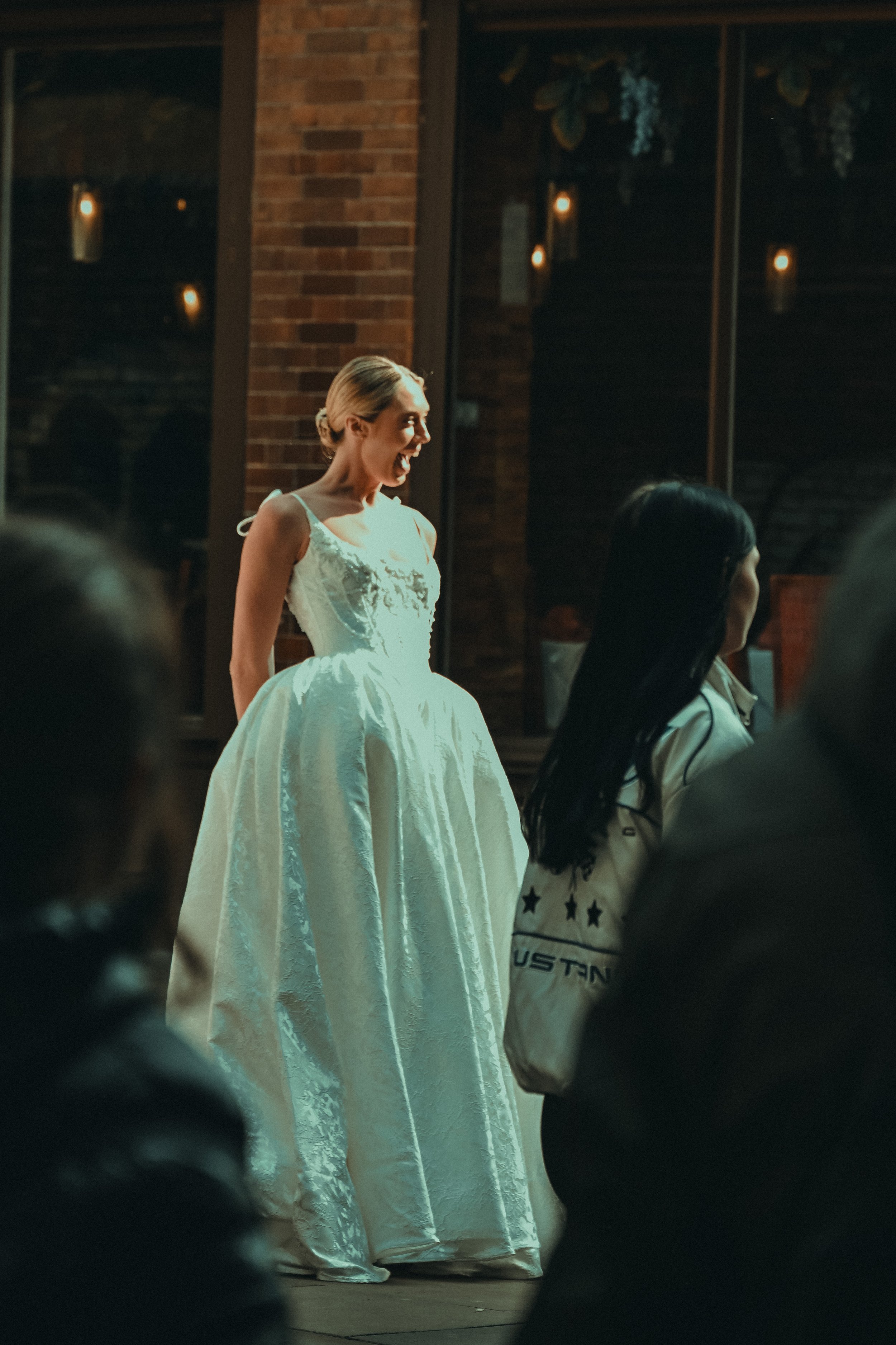 A woman wearing a white dress with intricate embroidery, smiling, standing in a warmly lit indoor space with brick walls and hanging lights, surrounded by an audience.