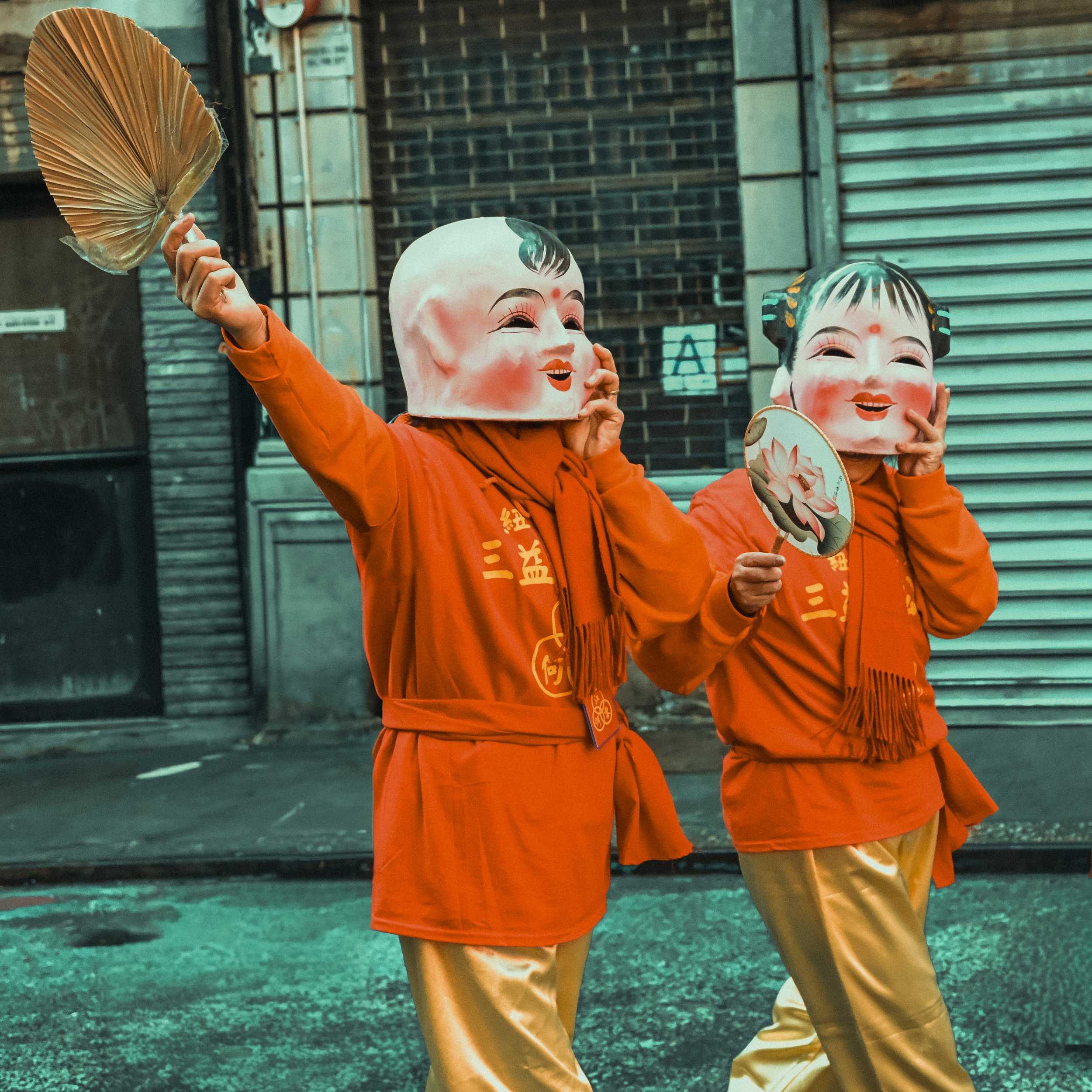 People wearing traditional Chinese opera masks and costumes, holding fans, walking on a street with a brick wall and metal shutter in the background.
