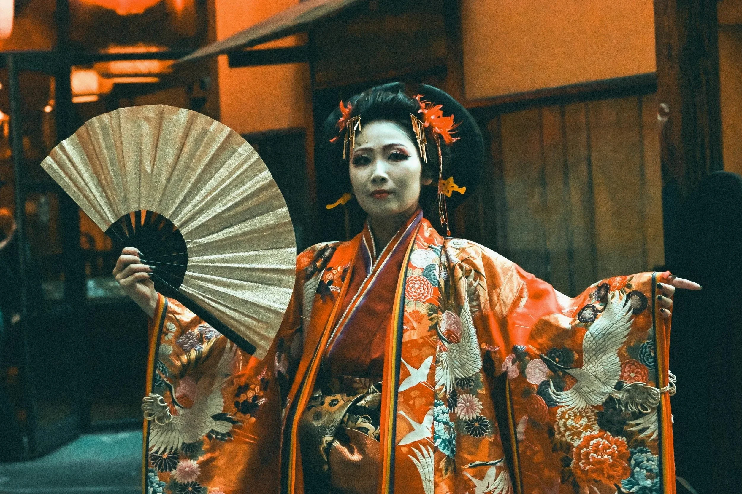 A person dressed in traditional Japanese geisha attire holding a folding fan with an intricate kimono and elaborate hairstyle, standing in a dimly lit indoor setting.