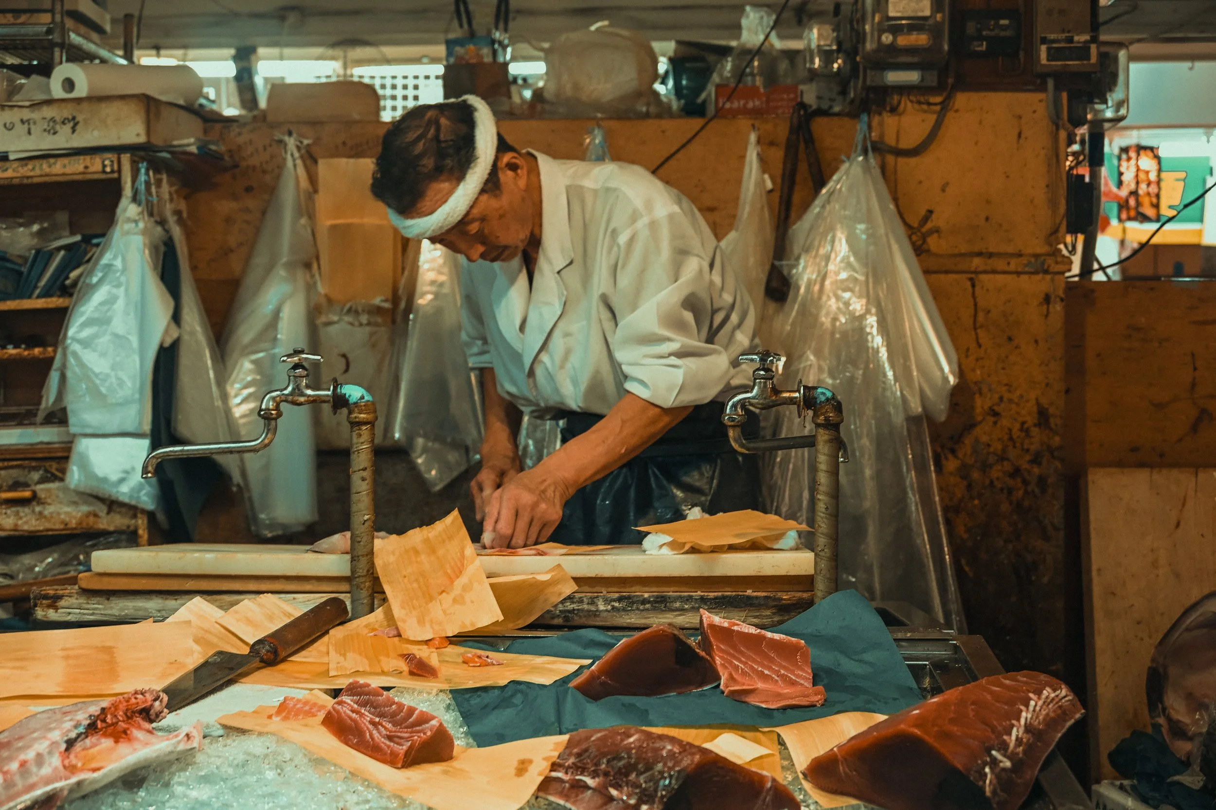 A man in a white shirt and headband prepares fish at a market stall with various fish cuts on the table.
