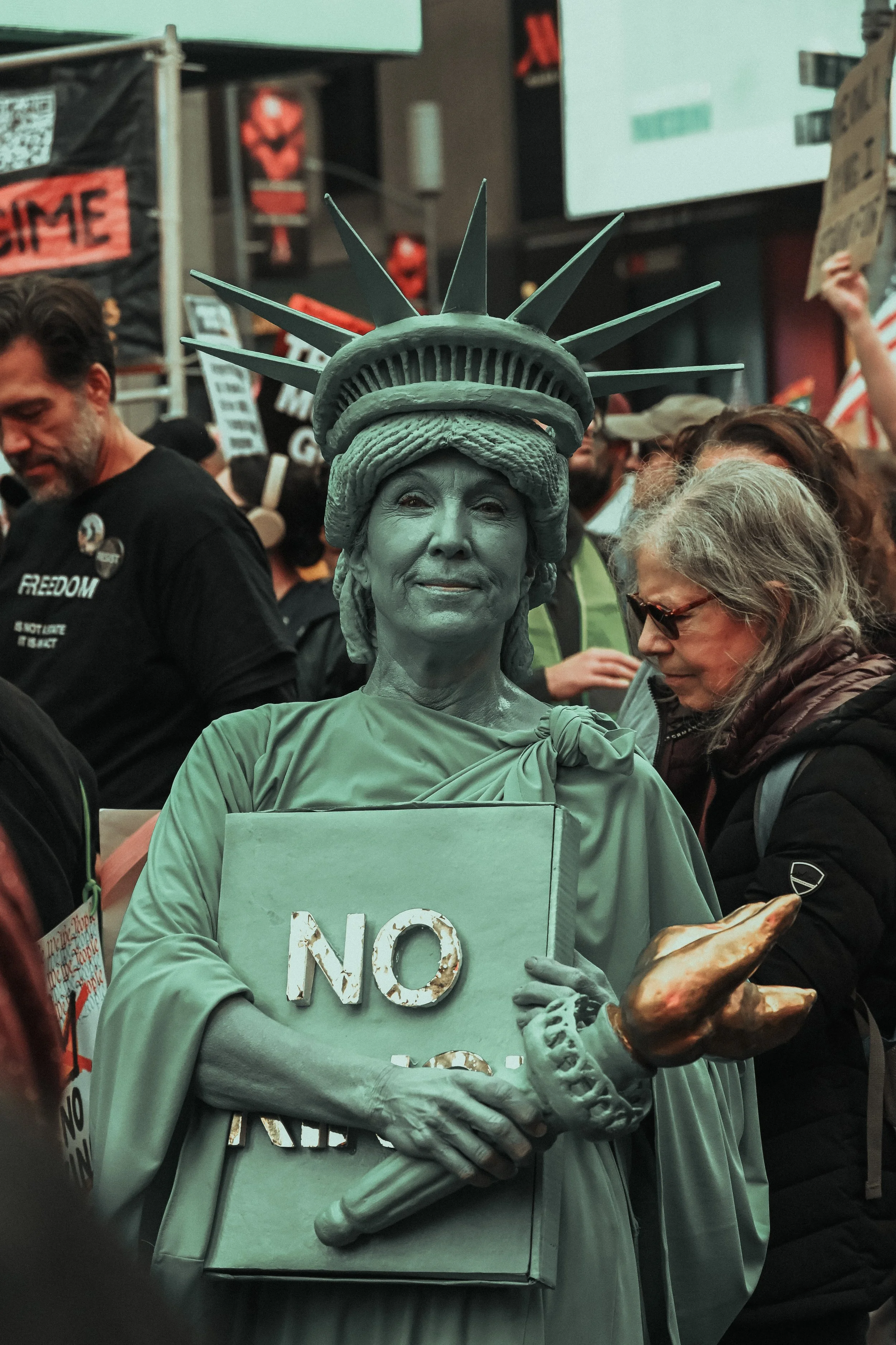 A woman dressed as the Statue of Liberty holding a sign that says "NO" at a protest or demonstration. She has a statue-like appearance with a crown and a greenish hue.