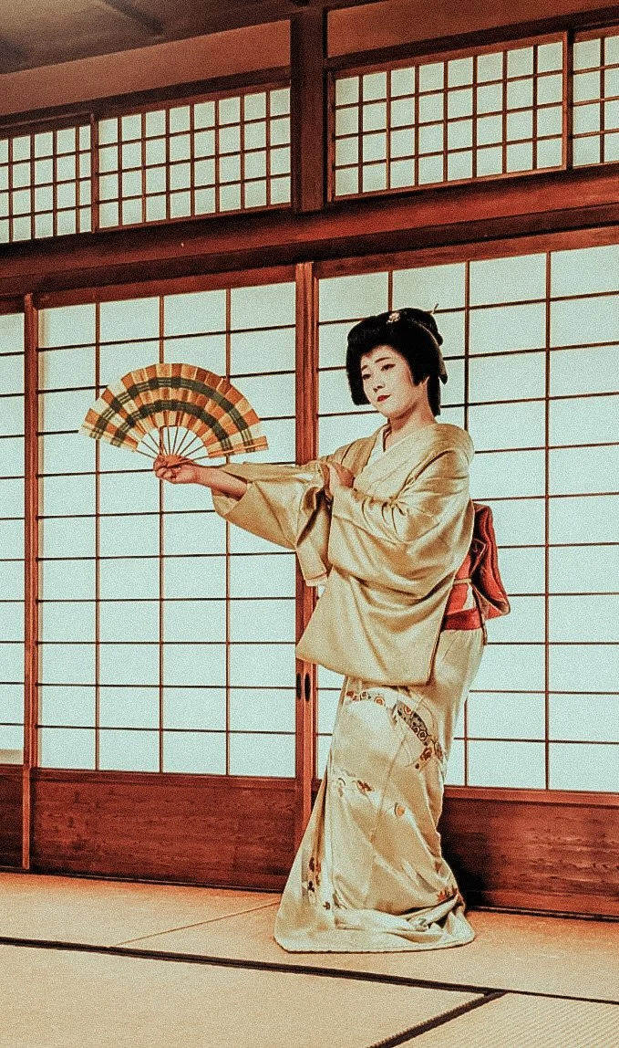 A woman in traditional Japanese kimono holding a folding fan in a room with shoji sliding doors.
