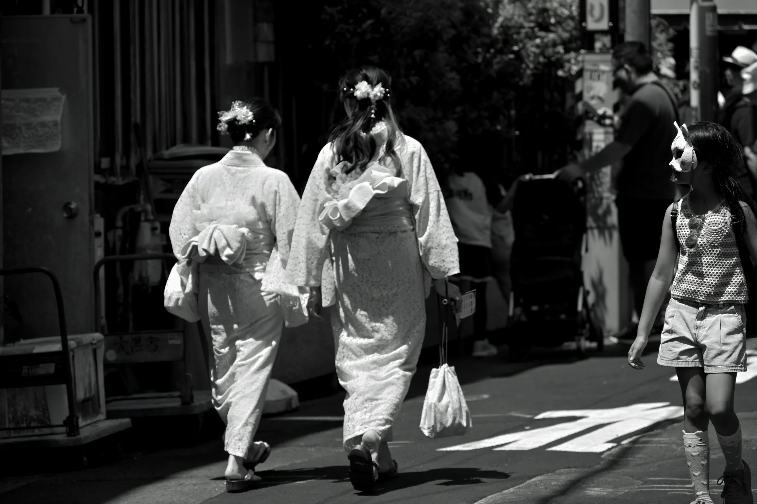 Three women in traditional Japanese kimonos walking together on a city street. Two women are facing away, and one woman is facing to the side, wearing a mask. Other pedestrians and street elements are visible in the background.