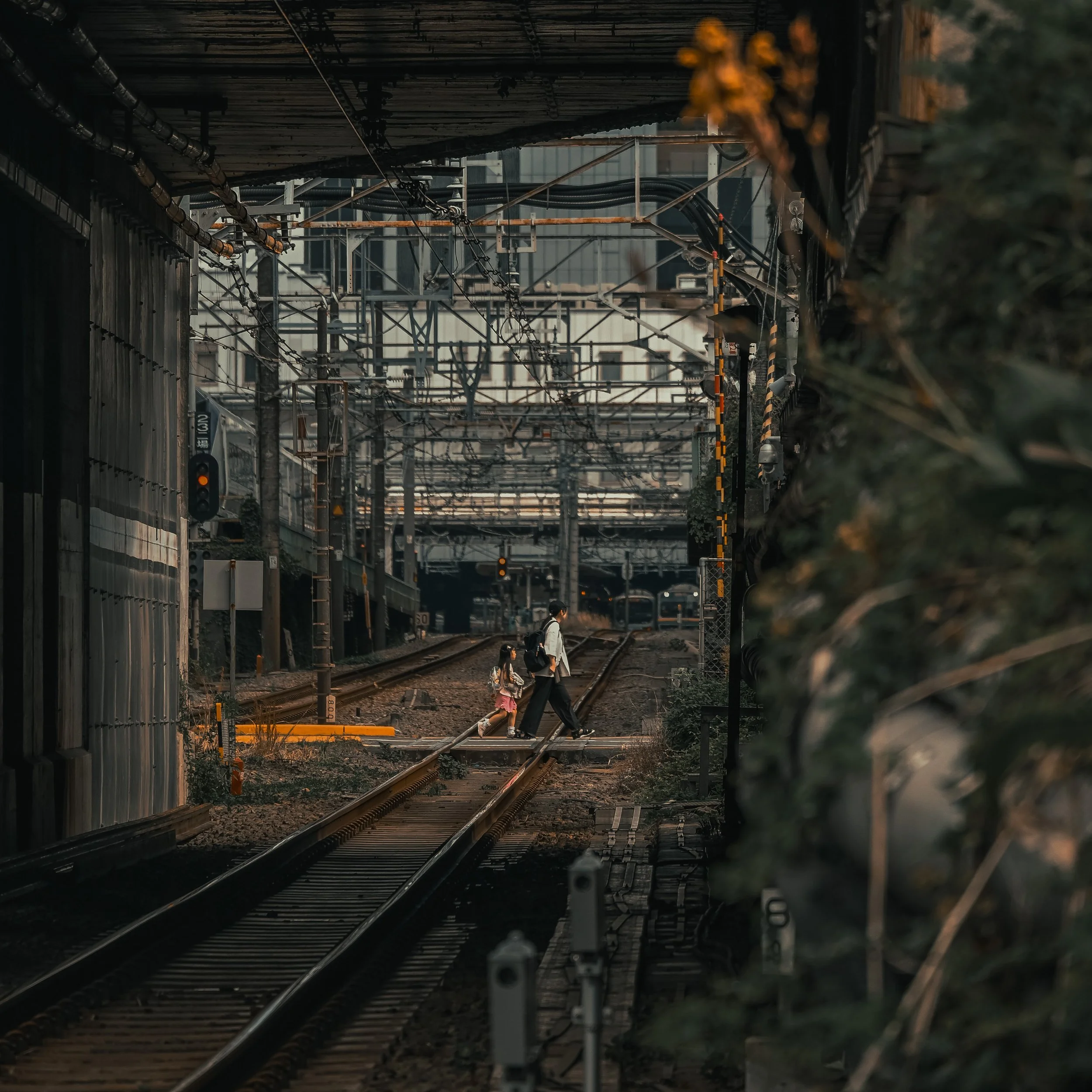 People crossing railway tracks under overhead electrical wires in an urban setting, with a train passing nearby and buildings in the background.