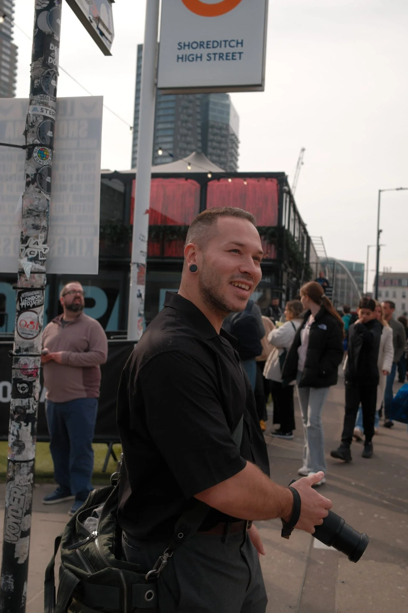 A smiling man with short hair and stretched earlobes holding a camera with a telephoto lens at an outdoor event in an urban setting. In the background, there are people, tall buildings, and a sign that reads 'Shoreditch High Street'.