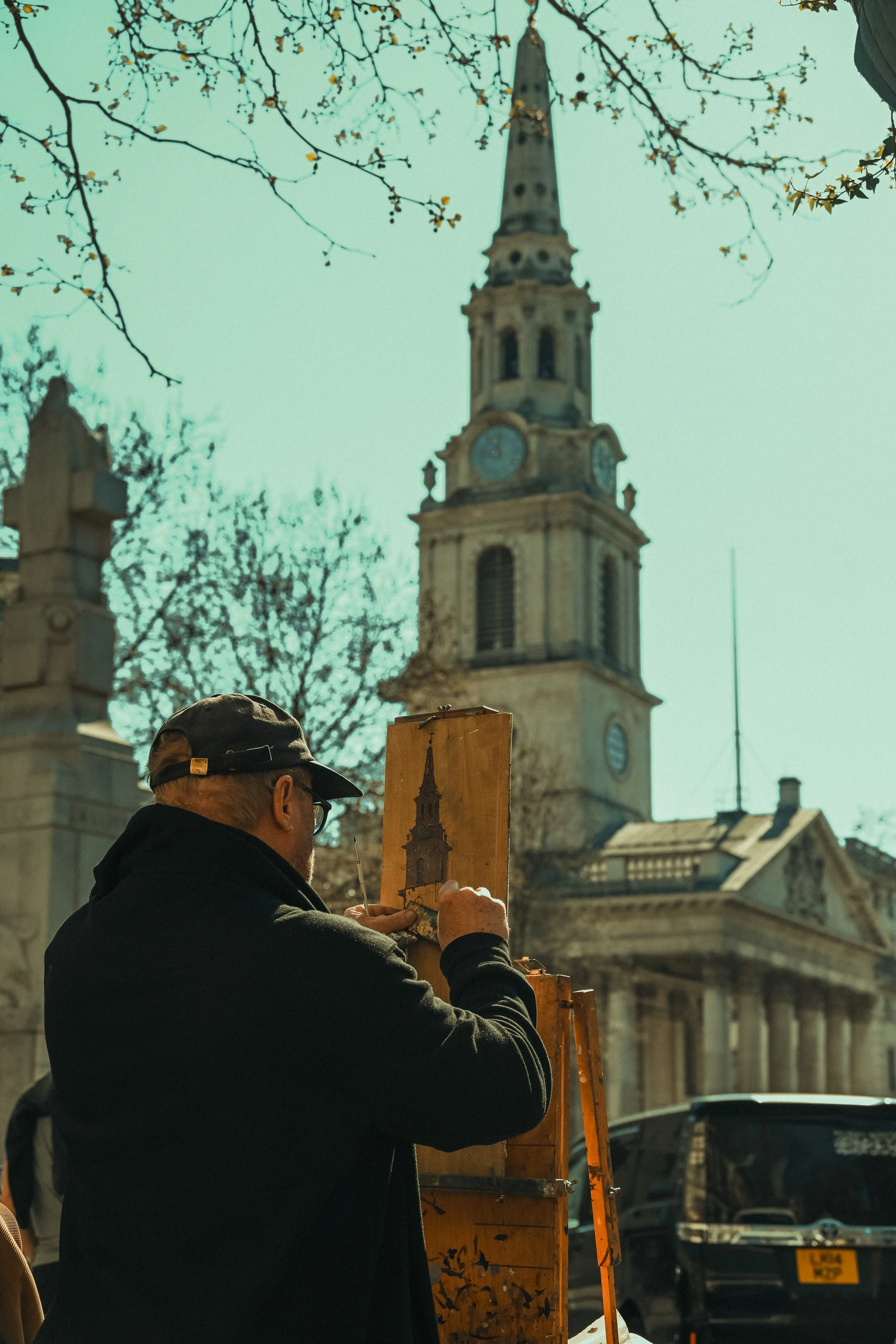 A man with a black cap and glasses painting a picture outdoors with a historical church tower and leafless trees in the background.