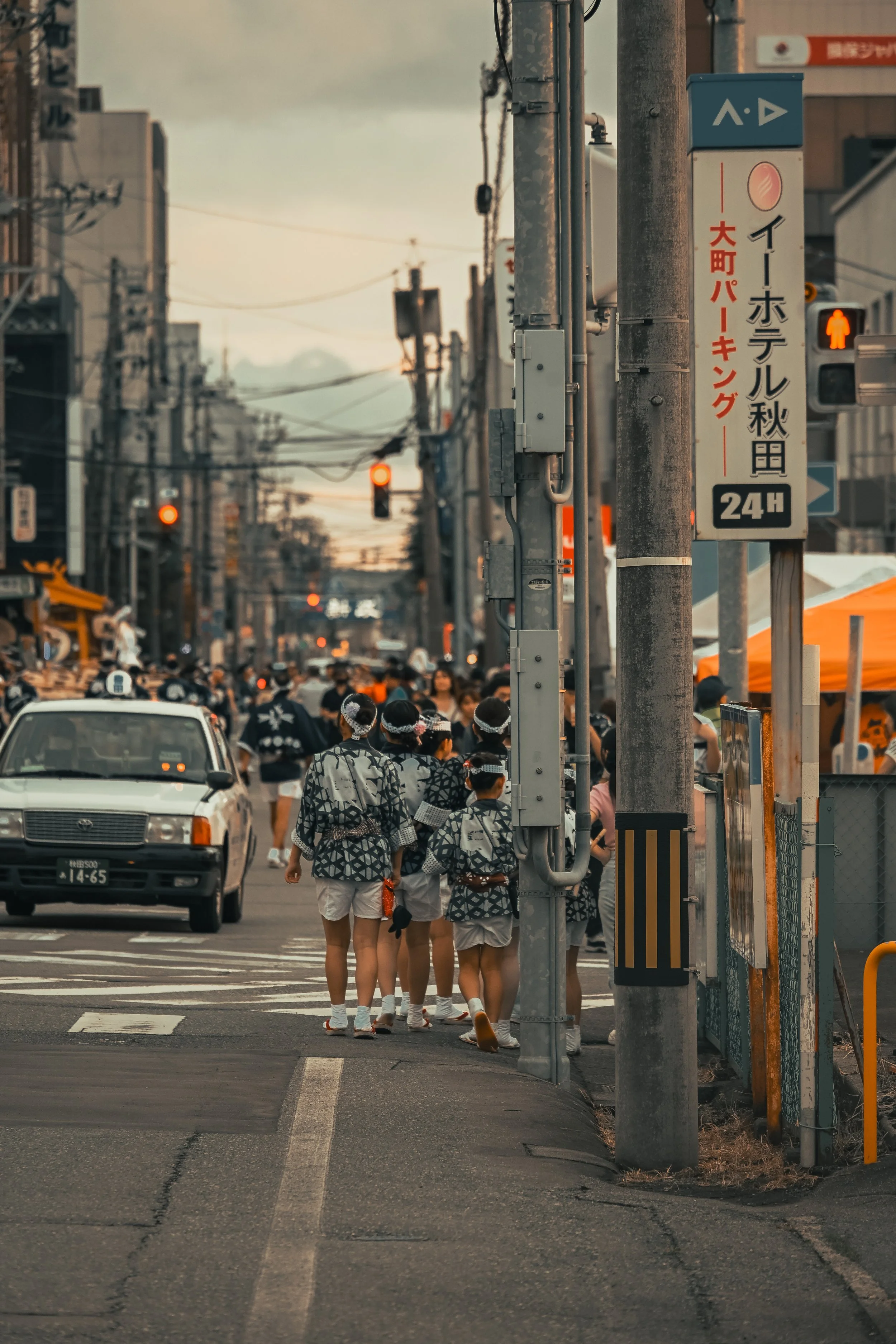 A group of children dressed in traditional Japanese festival clothing walking across a pedestrian crossing in an urban area during the evening or late afternoon, with Japanese signs and traffic lights visible.