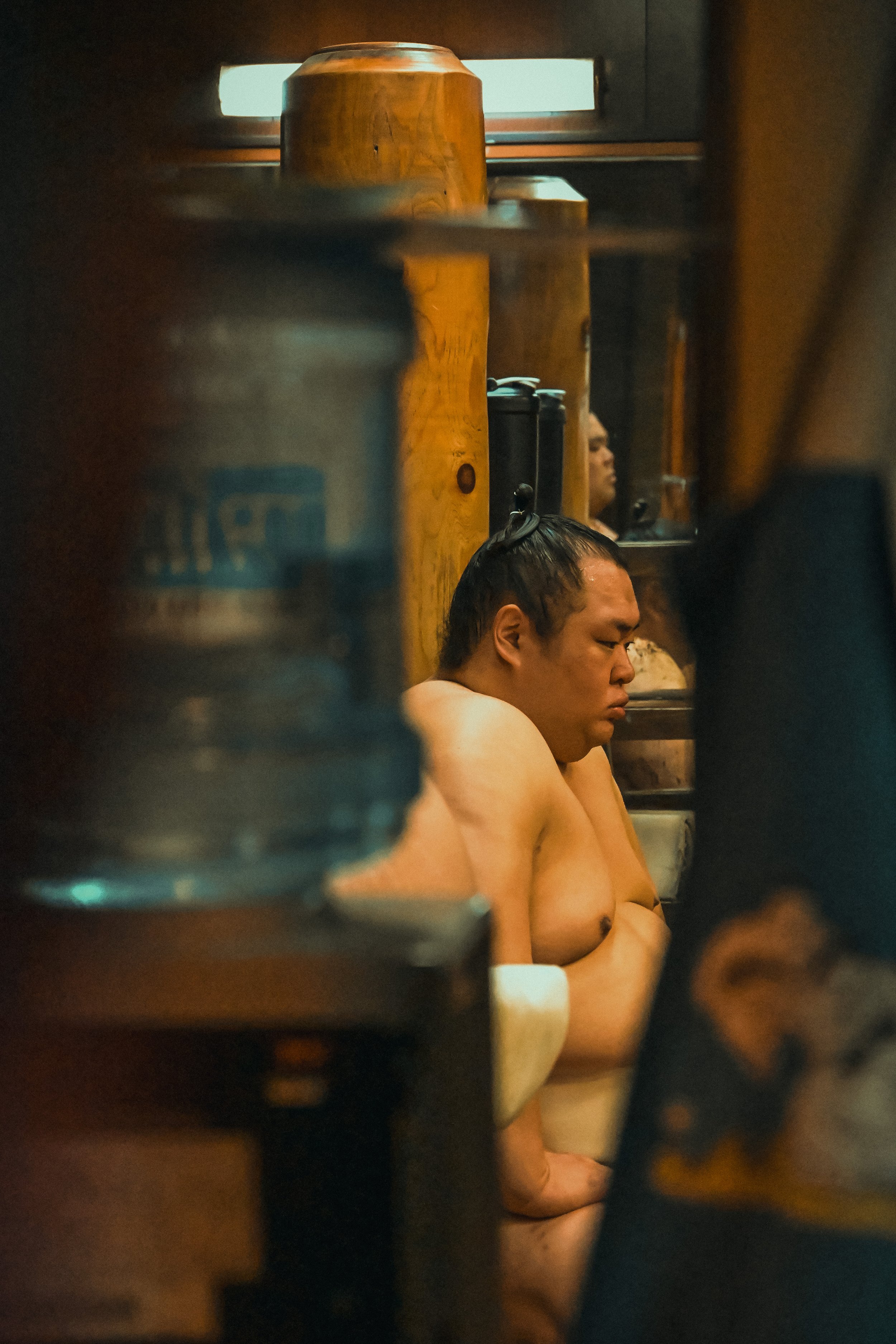 A man with long dark hair in a bun, shirtless, sitting in a traditional Japanese wooden bath or onsen, with a partially visible person in the background.
