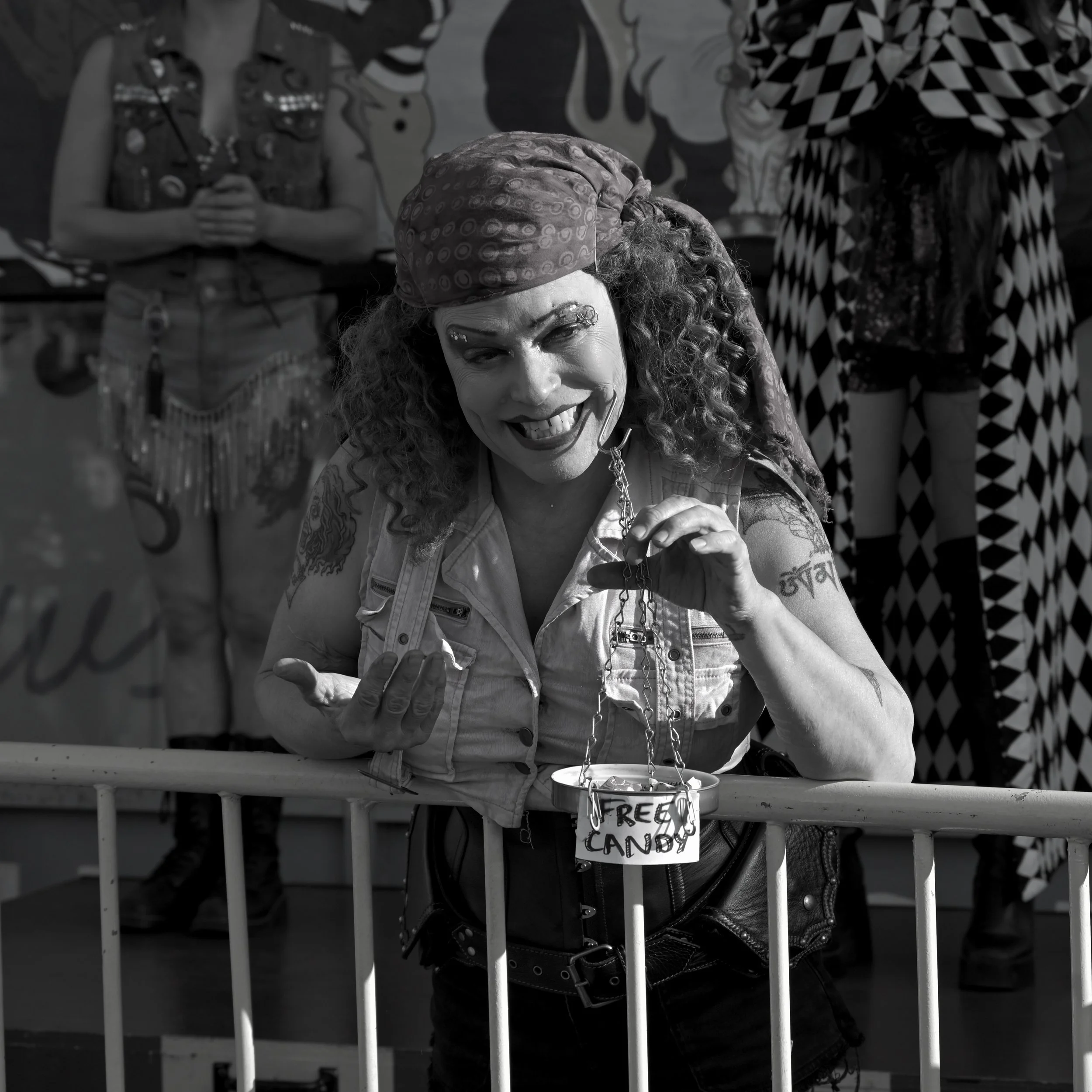 A woman with curly hair and a patterned headscarf smiling, holding a cane. Behind her, there are people in casual clothing and a sign that says 'Free Candy' hanging from the fence in front of her.