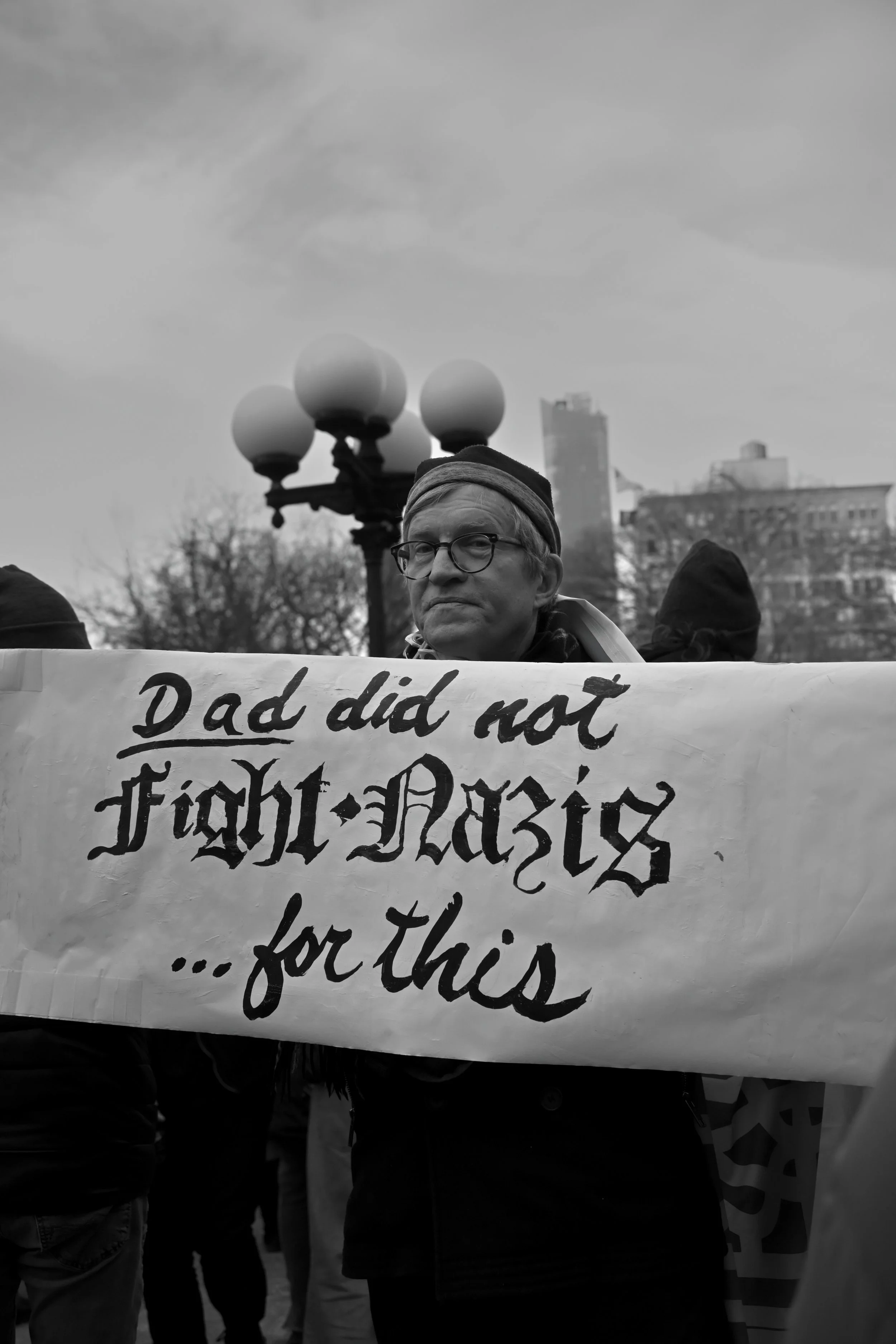 A man with glasses and a headband holds a protest sign with handwritten text. Other people are visible around him, and there are trees, a lamp post with multiple globes, and buildings in the background on a cloudy day.