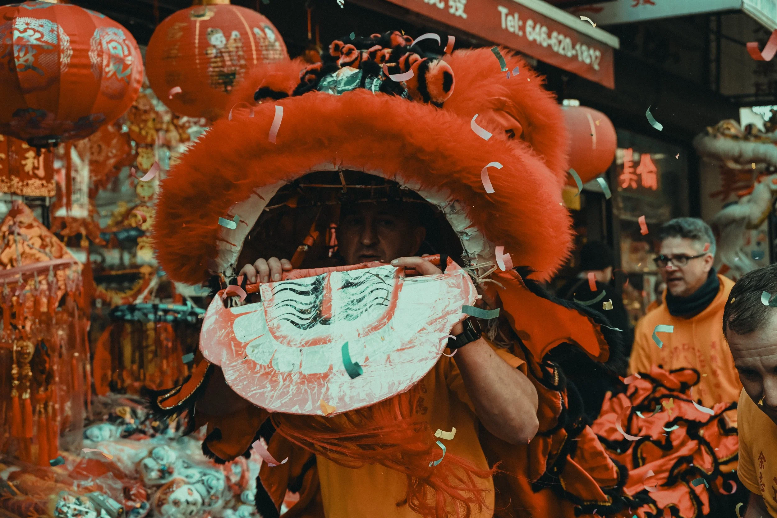 A man inside a red lion dance costume, emerging through the lion's head, during a celebration with red lanterns and decorations, and confetti in the air.