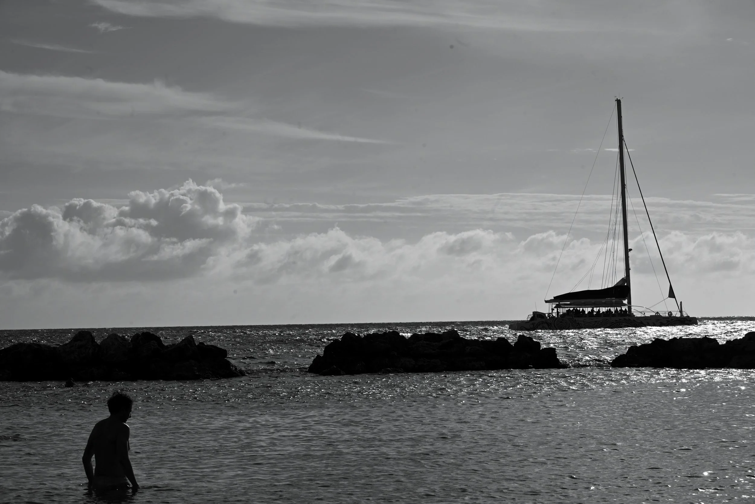A black-and-white photo of a person standing in the ocean near rocks with a sailboat in the background, clouds in the sky, and the sun reflecting on the water.