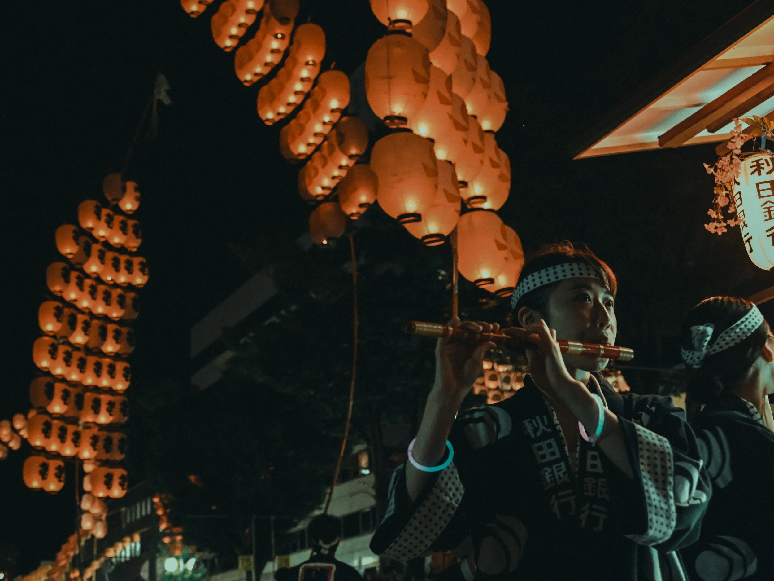 Children in traditional Japanese attire participating in a nighttime festival, holding small wooden flutes, with lanterns hanging overhead illuminating the scene.