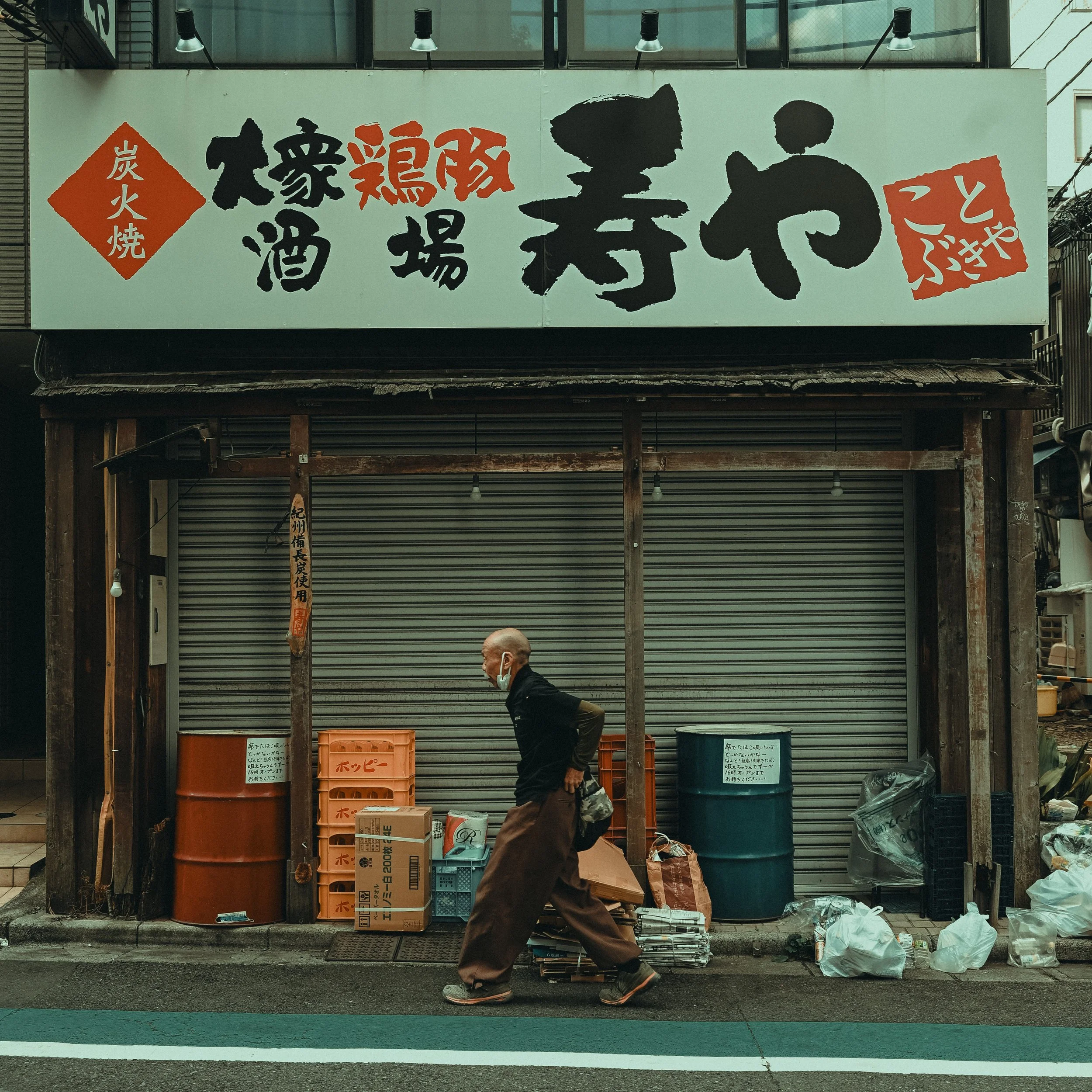 A man walking past a closed store with a large sign featuring Japanese characters. There are large barrels, boxes, and plastic bags with items on the ground in front of the store. The man is wearing a face mask and dark clothing.