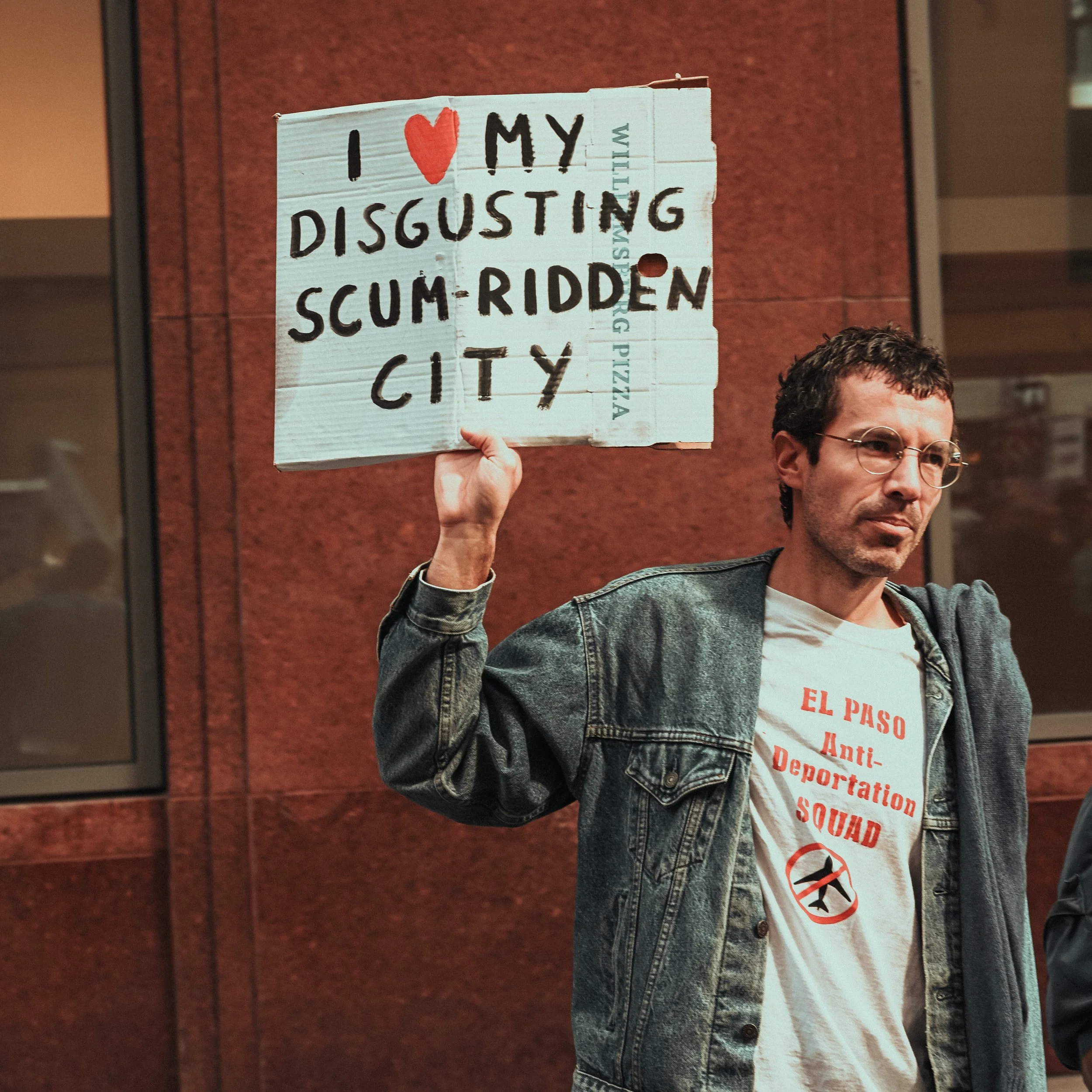 A protestor holding a cardboard sign that reads "I ❤️ MY DISGUSTING SCUM-RIDDEN CITY" in black and red text. The protestor is wearing glasses and a denim jacket, and there is a red brick wall background.